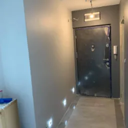 Korytarz w trakcie remontu z pomalowanymi na szaro ścianami, zabezpieczonymi drzwiami wejściowymi folią malarską i wbudowanymi w ścianę lampami LED.