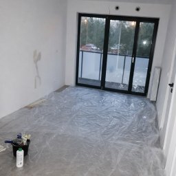 Radosław Majdanik - Usługi wykończeniowe - Przygotowanie do malowania: pokój zabezpieczony folią malarską, widoczne narzędzia (pędzle, wałki) i wiadro z farbą. Ściany częściowo zagruntowane, duże okno balkonowe.