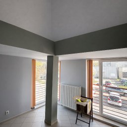 Radosław Majdanik - Usługi wykończeniowe - Jasne, odświeżone wnętrze po malowaniu: szare ściany, biały sufit z szarymi belkami, okna z pomarańczowymi żaluzjami. Karton na stojaku w rogu pokoju.