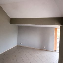 Radosław Majdanik - Usługi wykończeniowe - Pomalowane na szaro puste wnętrze z beżową podłogą z płytek, widoczne gniazdka elektryczne i fragment okna z żaluzjami. Świeżo po remoncie.