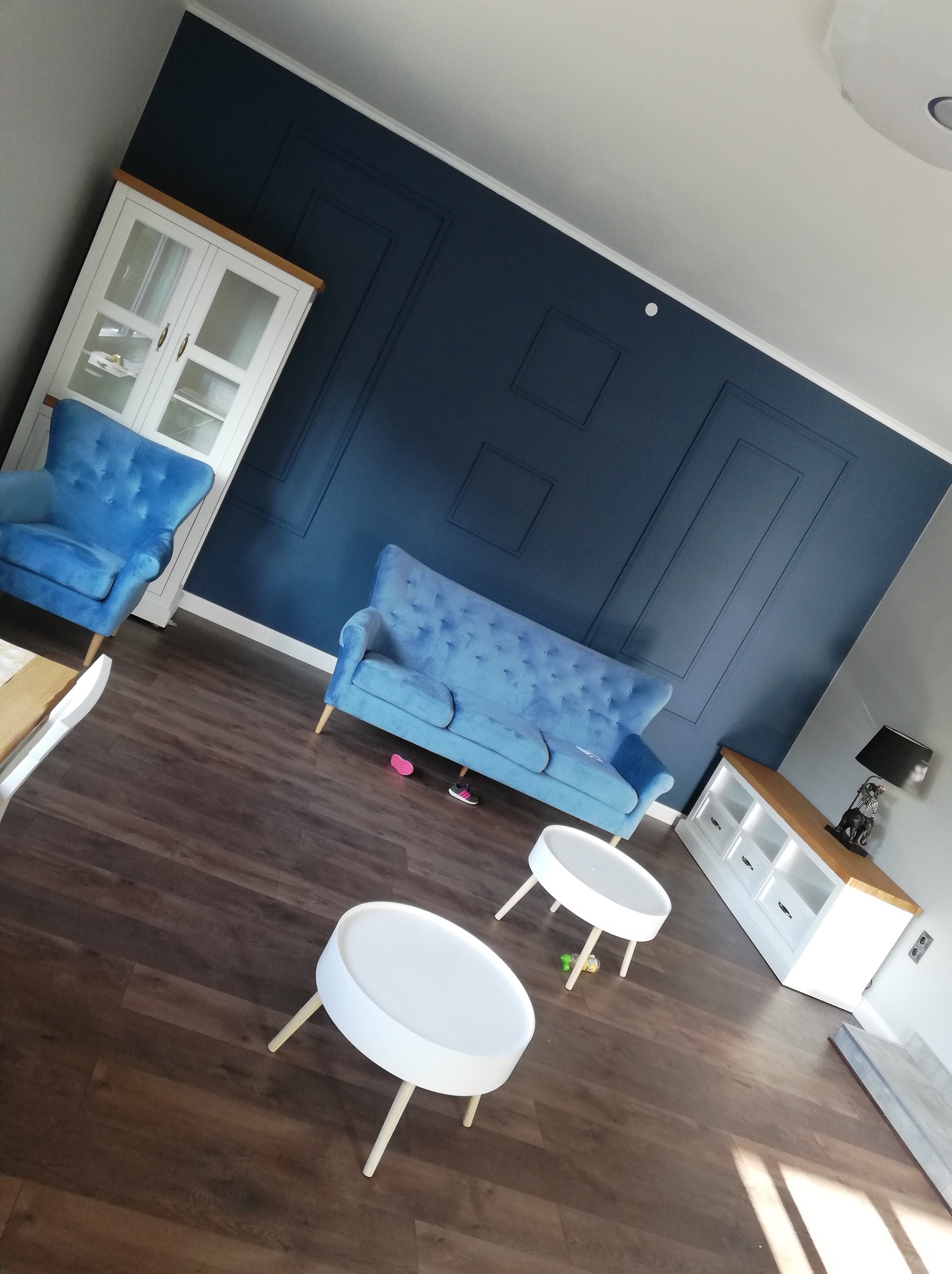 Salon z ciemną, dekoracyjną ścianą, błękitnym, tapicerowanym kompletem wypoczynkowym i białymi stolikami kawowymi na drewnianej podłodze.