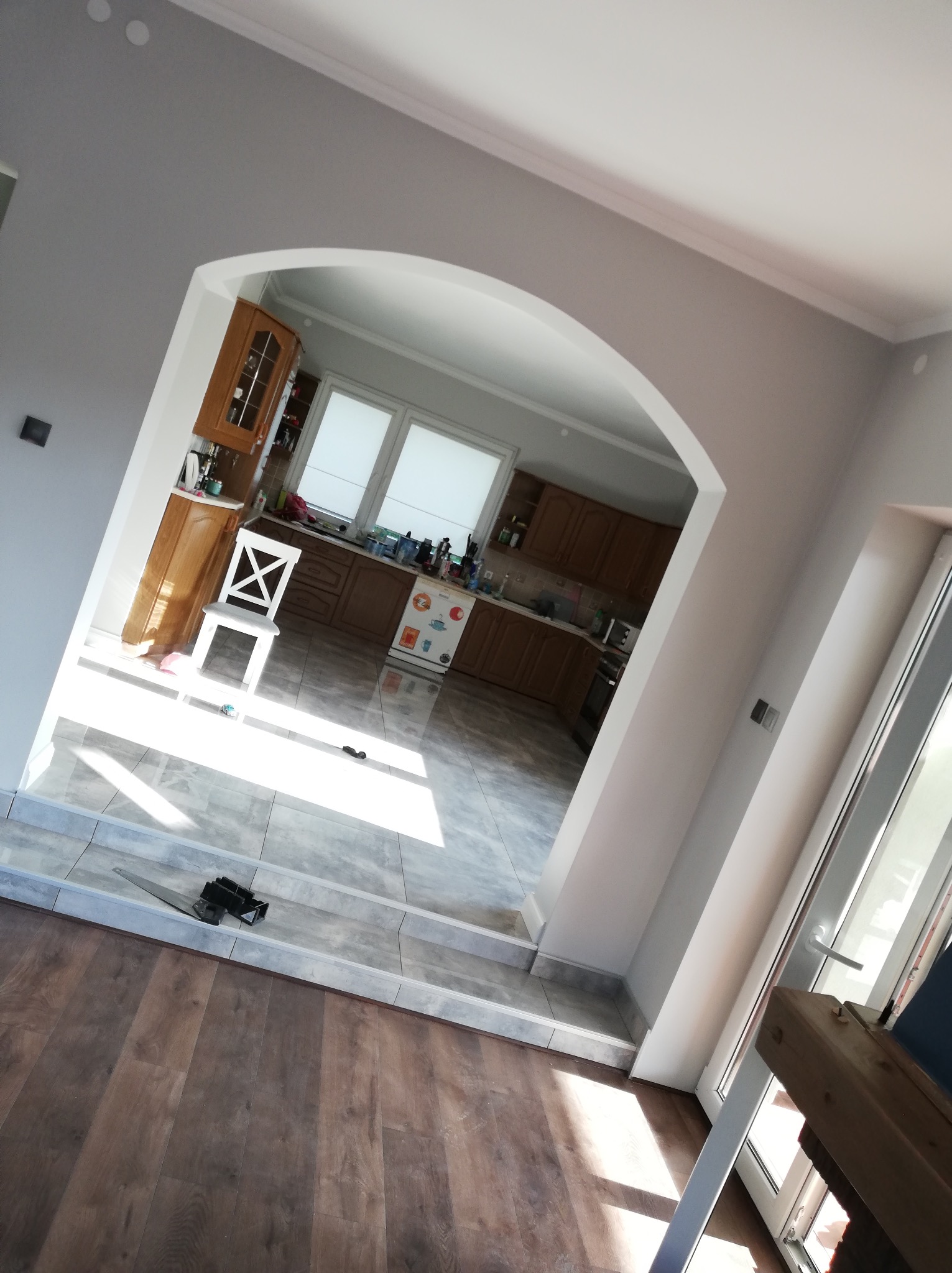 Arched doorway connecting a living room with dark wood flooring to a bright kitchen with gray tile flooring and white chair, freshly painted gray walls.