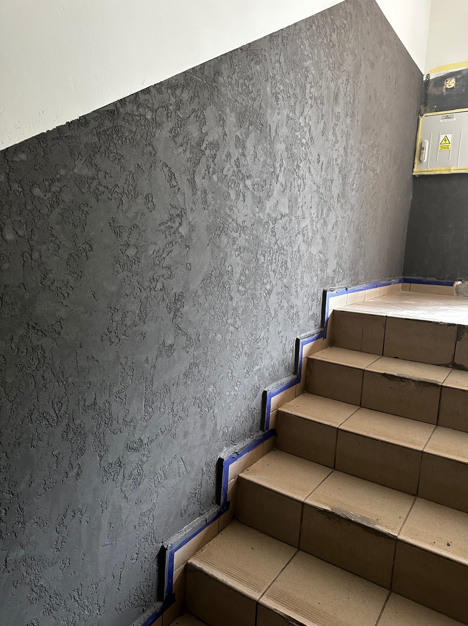 Klatka schodowa z szarą, teksturowaną ścianą, zabezpieczona niebieską taśmą malarską przy schodach z płytek. Widoczna skrzynka elektryczna na ścianie w tle.