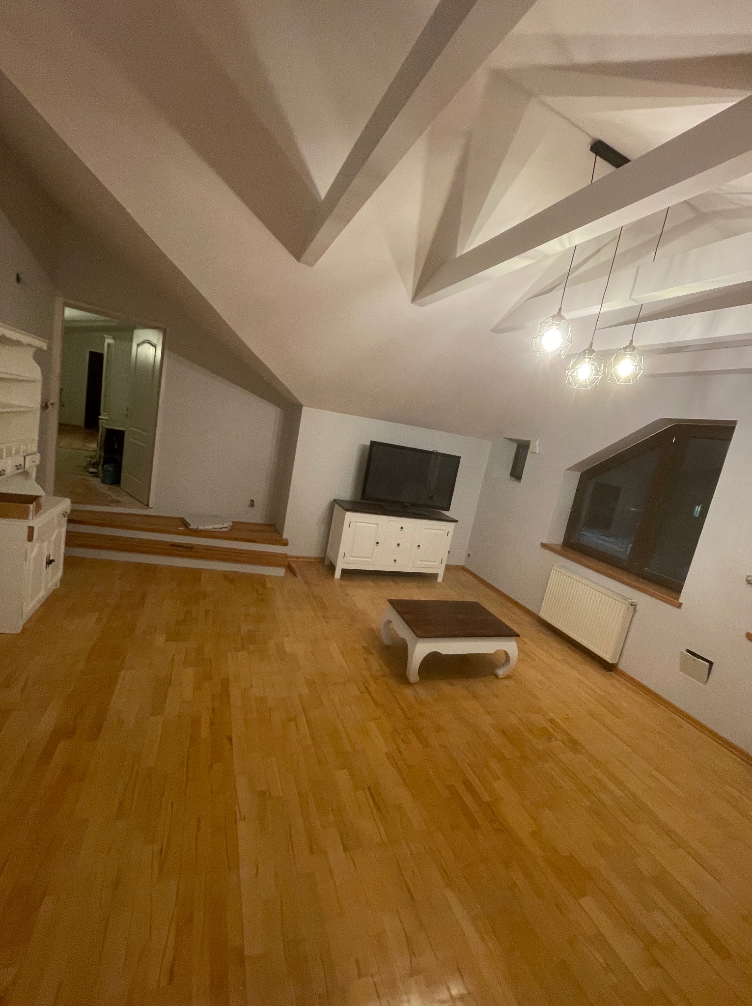 Jasne, przestronne poddasze po remoncie z drewnianą podłogą, białymi meblami i skośnym sufitem z widocznymi belkami.