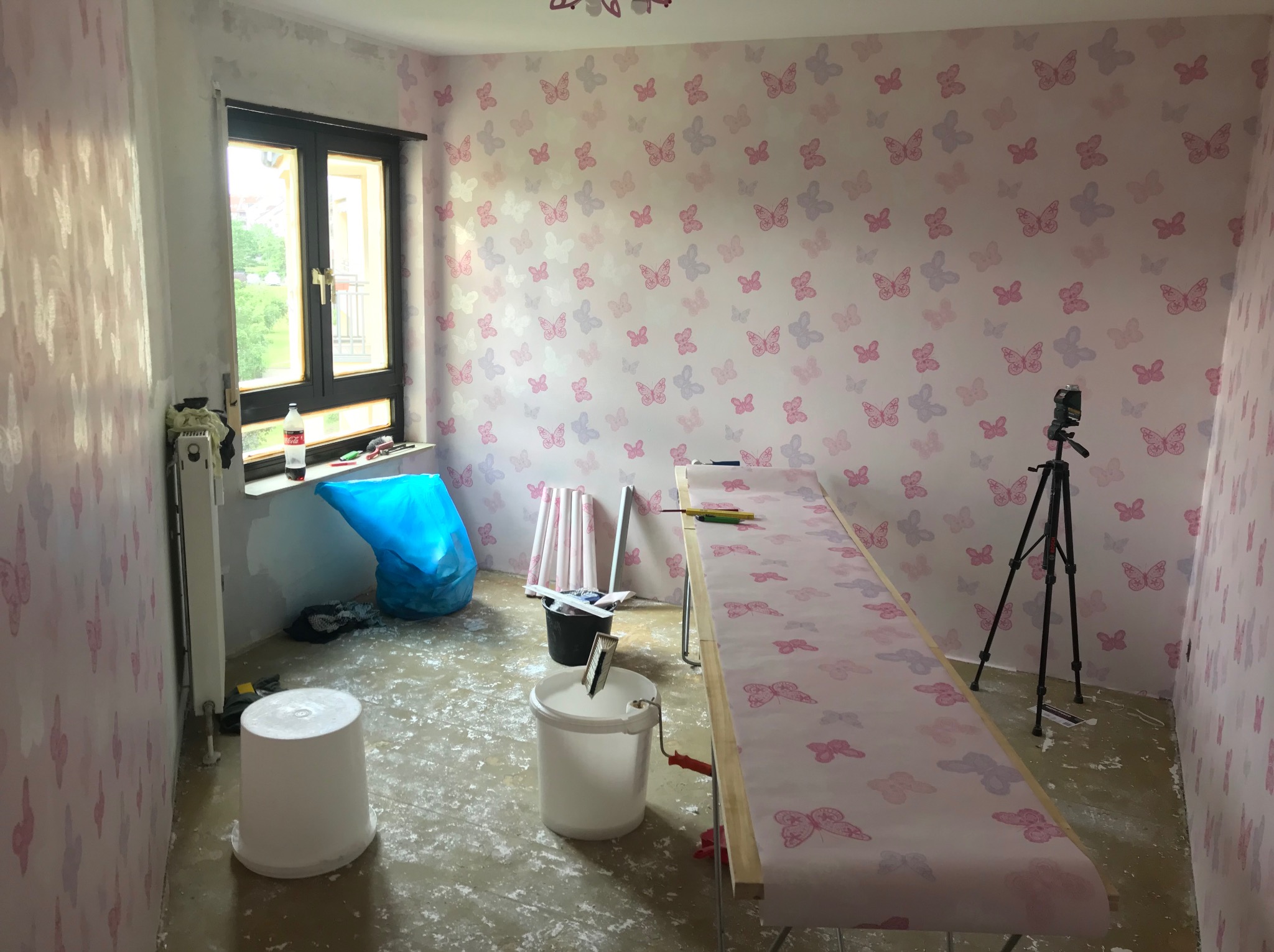 Pokój z motywem motyli w trakcie remontu: ściany pokryte tapetą w motyle, widoczne narzędzia malarskie, wiadro z farbą, stół roboczy z rozłożoną tapetą i laserowy niwelator na statywie.