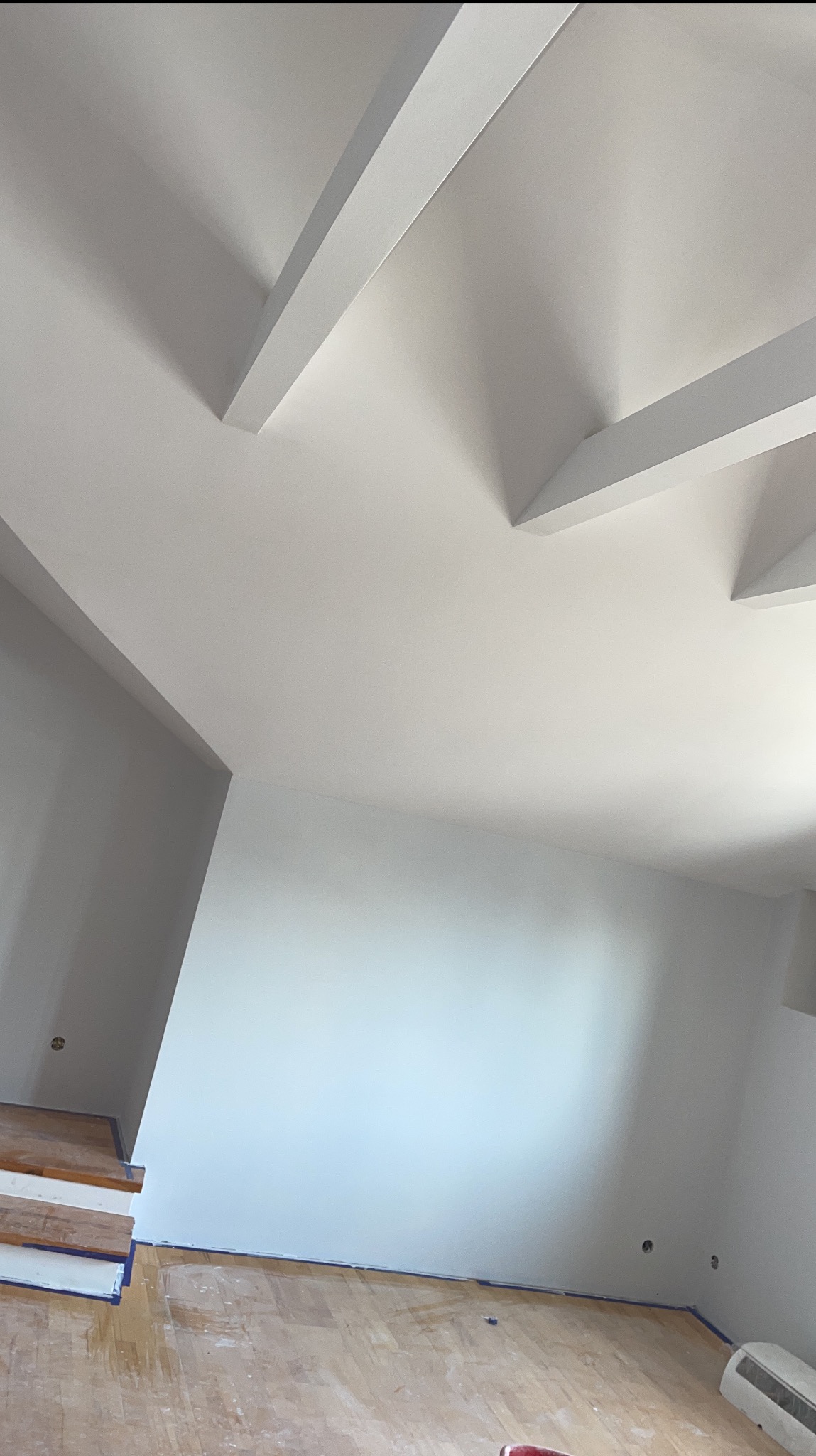 Świeżo pomalowane na biało wnętrze z drewnianą podłogą zabezpieczoną taśmą malarską, widoczne fragmenty grzejnika i schodów.