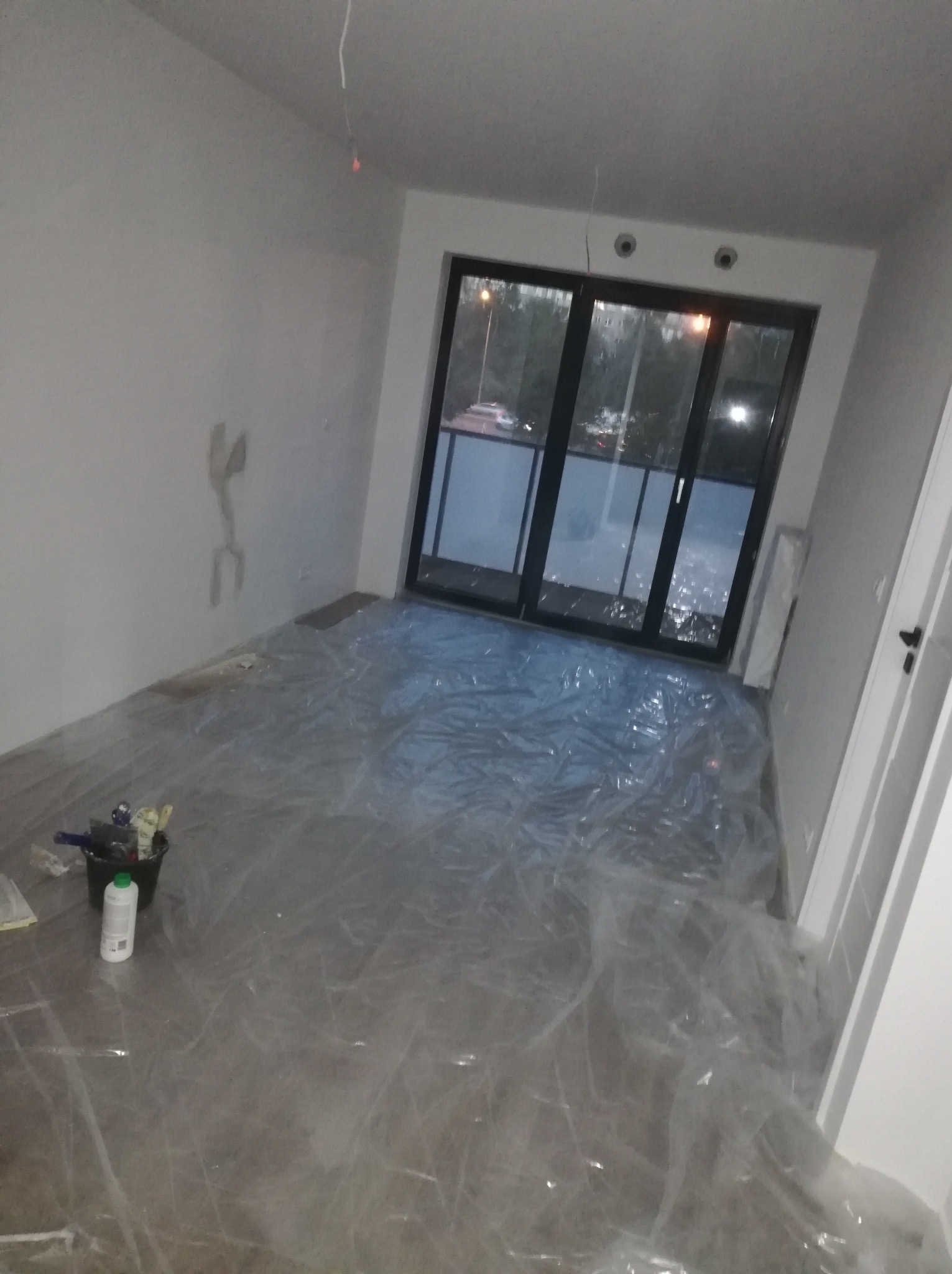 Przygotowanie pokoju do malowania: podłoga zabezpieczona folią malarską, widoczne narzędzia i pojemnik z farbą, ściany z miejscowymi naprawami, okno balkonowe.