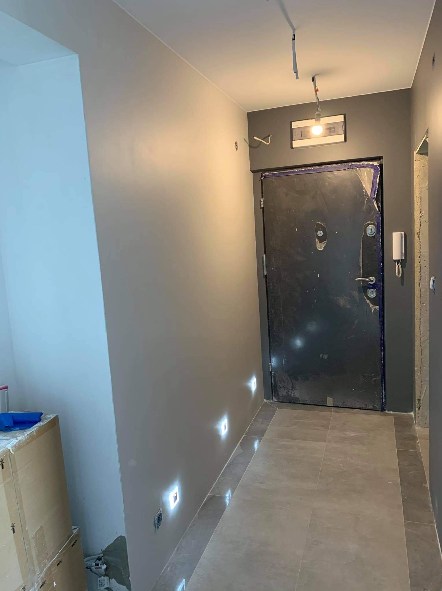 Korytarz w trakcie remontu z pomalowanymi na szaro ścianami, zabezpieczonymi drzwiami wejściowymi folią malarską i wbudowanymi w ścianę lampami LED.