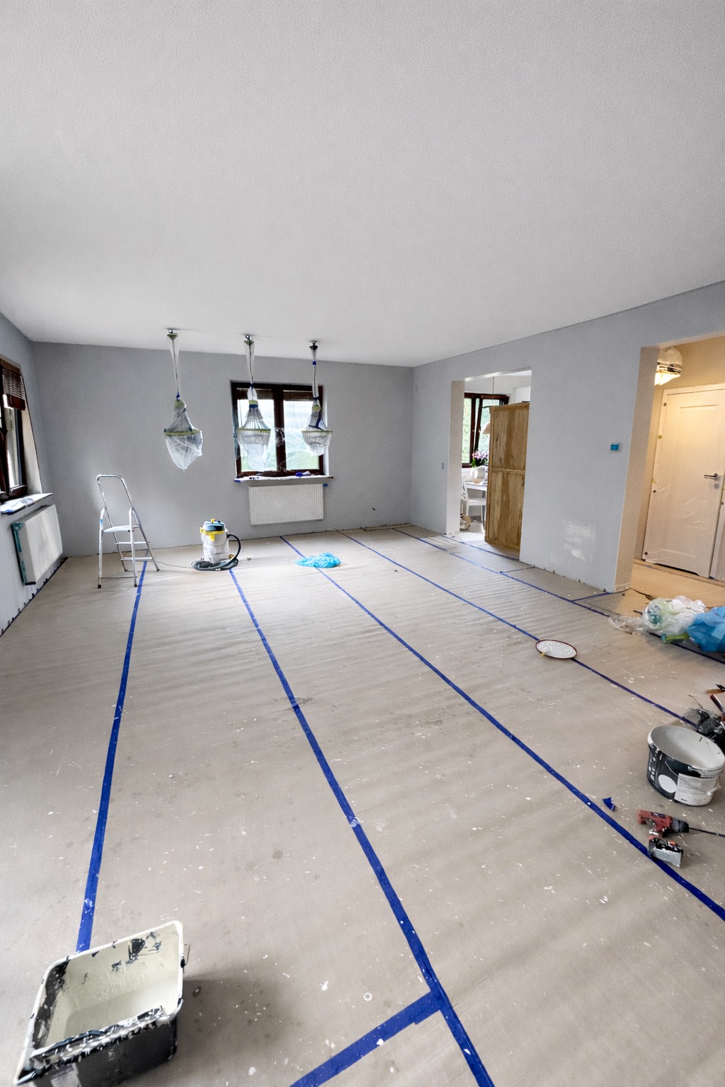 Przygotowane do malowania wnętrze: podłoga zabezpieczona folią i taśmą, ściany szare, okna z osłoniętymi lampami, widoczne narzędzia malarskie i drabina.