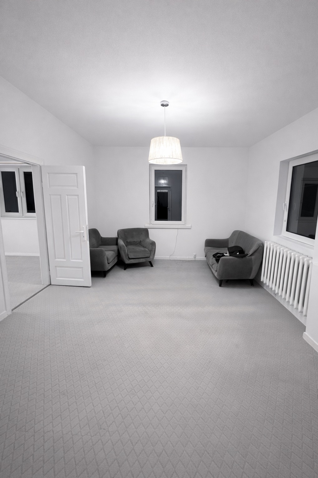 Jasne, przestronne wnętrze po remoncie: szare meble wypoczynkowe, białe ściany i grzejnik, wzorzysta wykładzina. Widok na sąsiednie pomieszczenie przez otwarte drzwi.