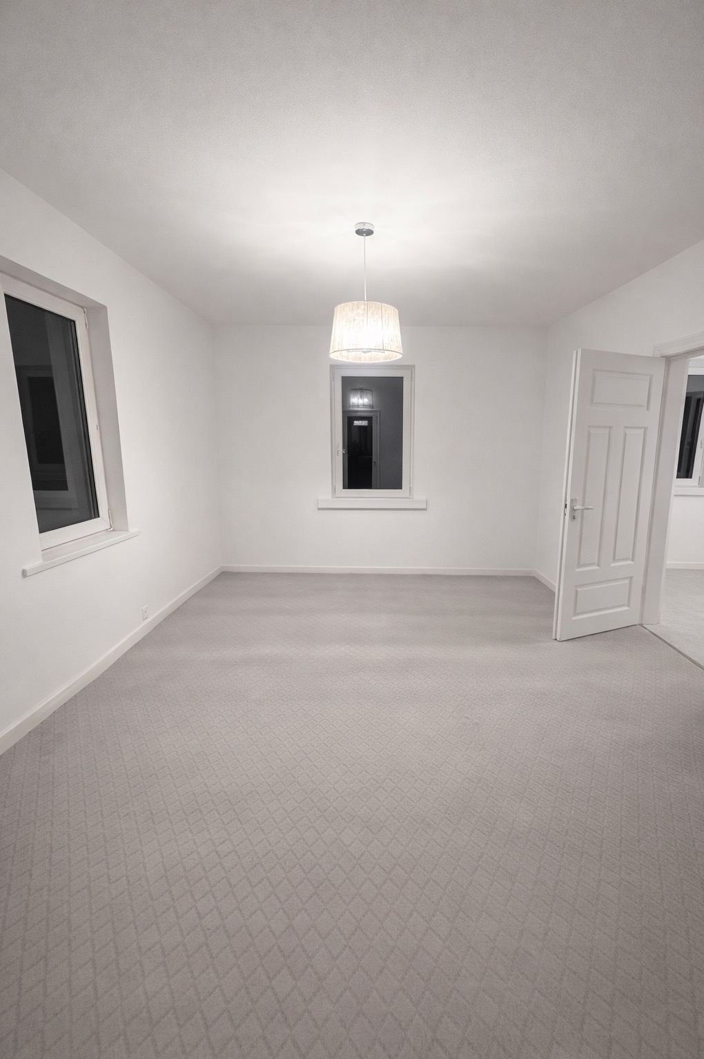 Jasne, puste wnętrze po remoncie: białe ściany, szara wykładzina w romby, okno z widokiem na zewnątrz i otwarte białe drzwi. Centralnie lampa wisząca.
