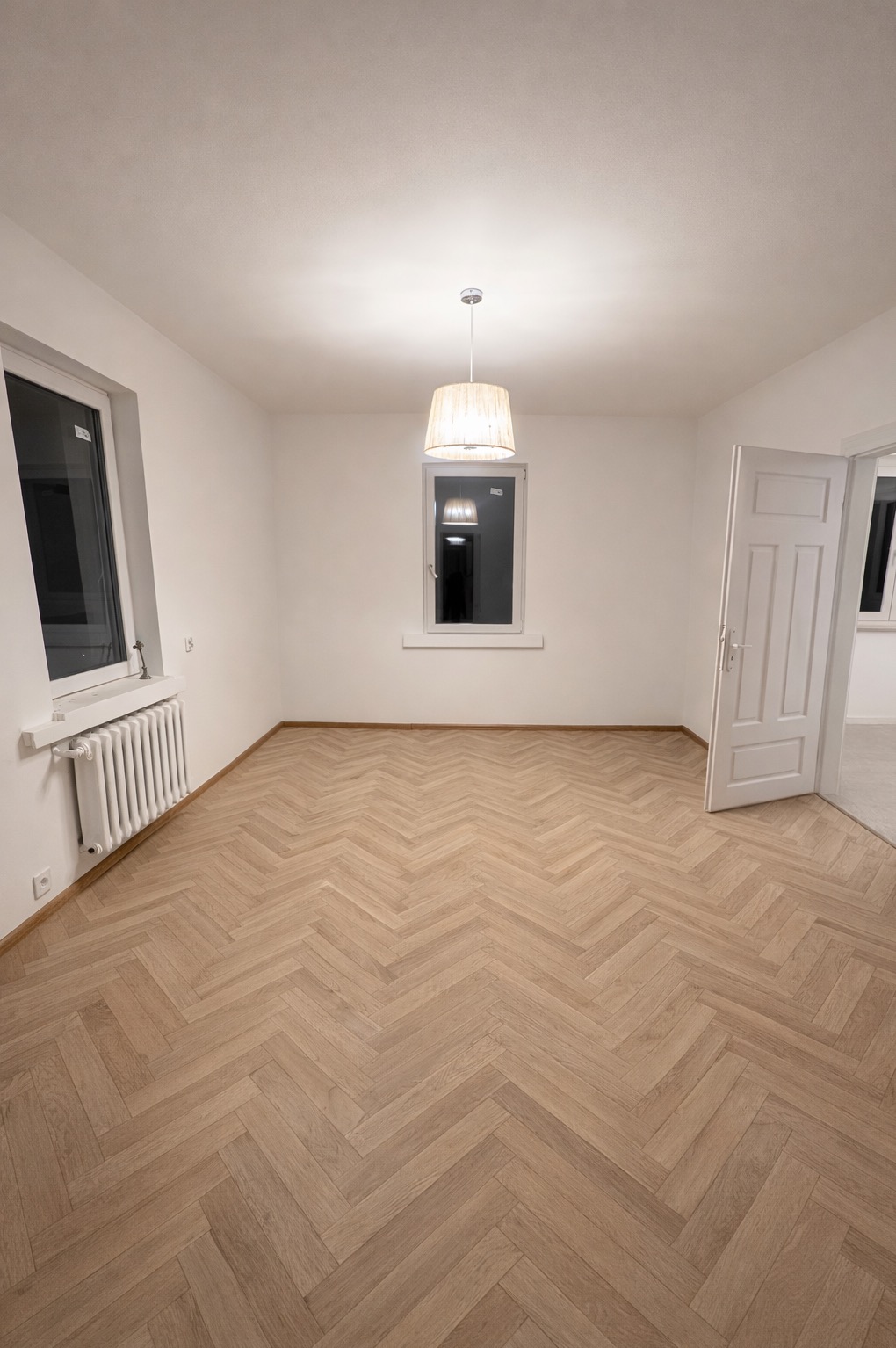 Pusty pokój po remoncie z parkietem w jodełkę, białymi ścianami, grzejnikiem i lampą wiszącą. Widok na otwarte drzwi do kolejnego pomieszczenia.