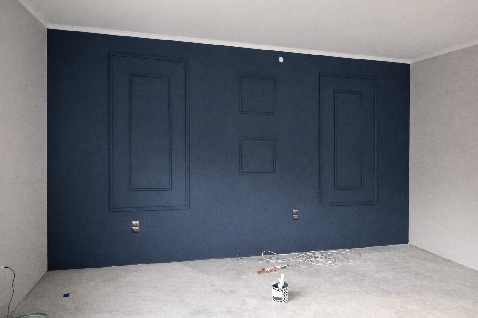 Granatowa ściana z dekoracyjnymi ramkami i listwami, świeżo pomalowana, z widocznymi gniazdkami i akcesoriami malarskimi na podłodze. Wykończenie wnętrza w trakcie realizacji.