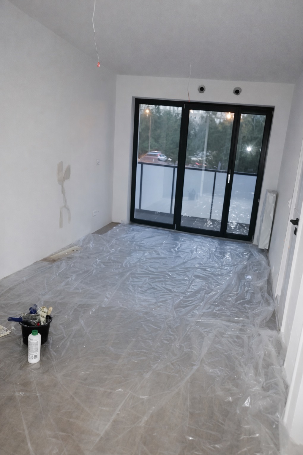 Przygotowanie do malowania: pokój zabezpieczony folią malarską, widoczne narzędzia (pędzle, wałki) i wiadro z farbą. Ściany częściowo zagruntowane, duże okno balkonowe.