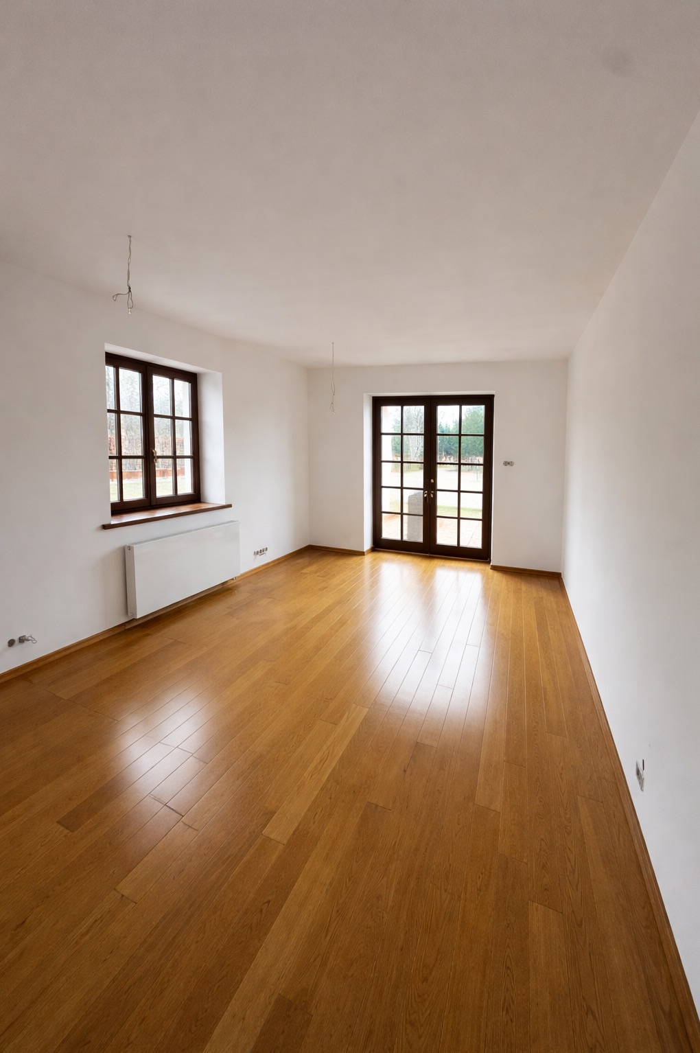 Jasne, puste wnętrze z drewnianą podłogą, białymi ścianami i oknem z grzejnikiem. Widoczne drzwi balkonowe z ciemnego drewna. Minimalistyczny design, świeżo po remoncie.