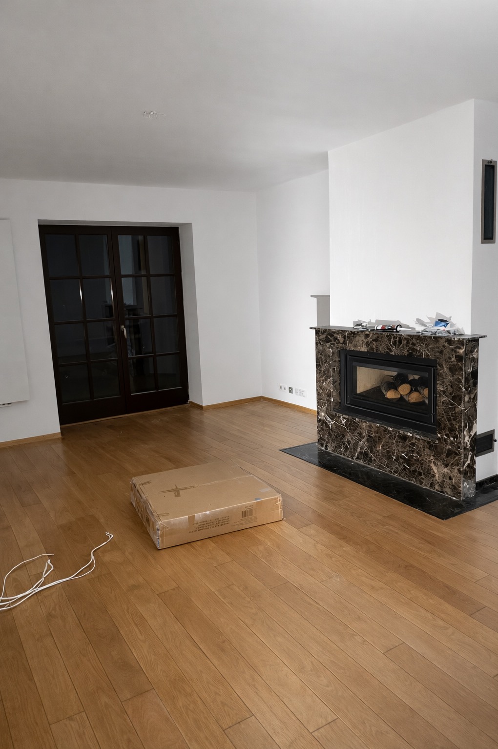 Jasne, puste wnętrze z drewnianą podłogą i marmurowym kominkiem. Na podłodze leży zapakowany karton i biały kabel. Widoczne dwuskrzydłowe drzwi balkonowe.
