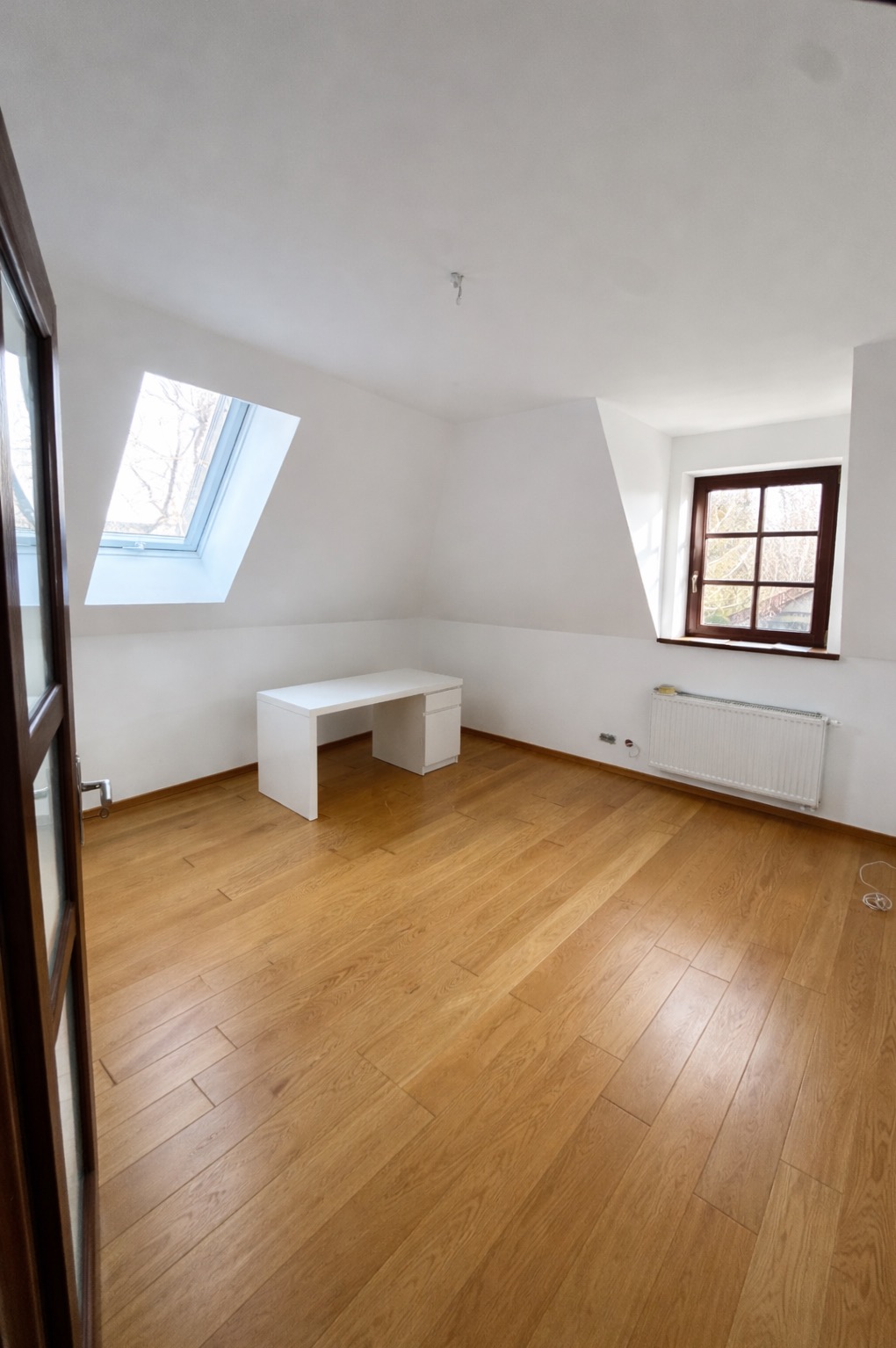 Jasne, puste poddasze z drewnianą podłogą, białymi ścianami i oknami dachowymi. Widoczny biały biurko i grzejnik. Ujęcie z perspektywy wejścia do pokoju.