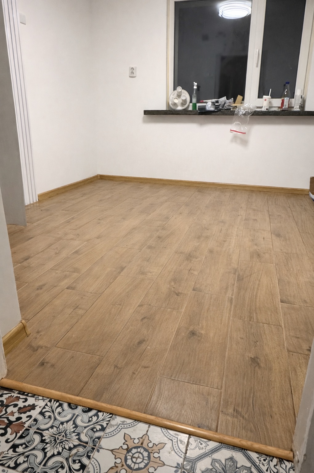 Ukończona podłoga z paneli imitujących drewno w jasnym pomieszczeniu. Widoczne listwy przypodłogowe i przejście do pomieszczenia z wzorzystą podłogą.