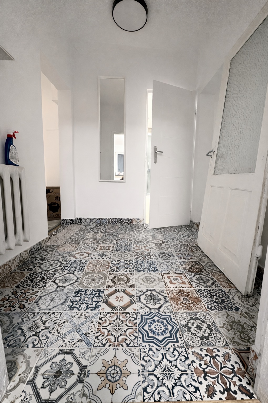 Białe wnętrze po renowacji z wzorzystą, kolorową podłogą z płytek. Widoczne drzwi, lustro i fragment grzejnika. Całość w jasnej tonacji.