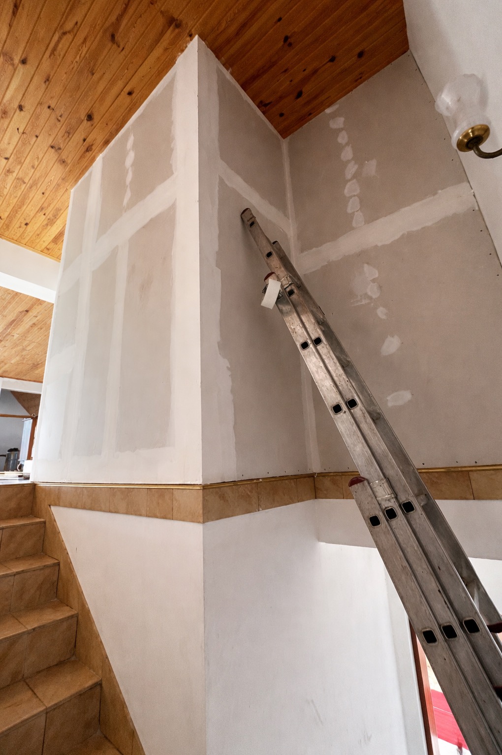 Ściana po szpachlowaniu, z widocznymi łączeniami płyt gipsowych i drabiną. Wykończenie sufitu boazerią. Prace remontowe wewnątrz budynku.