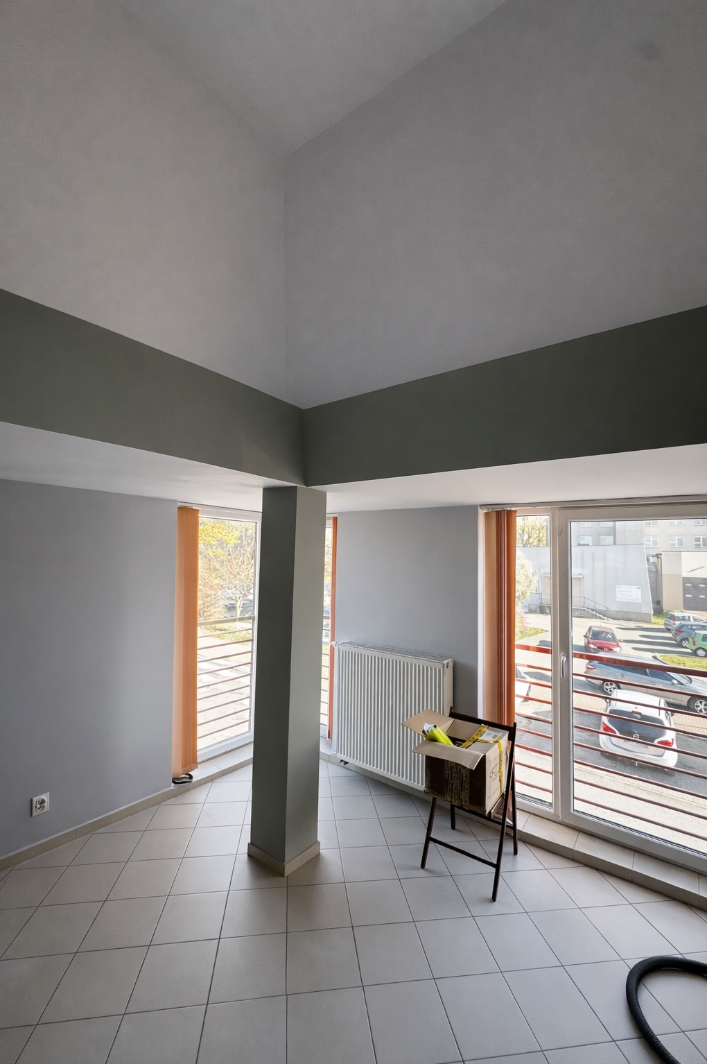 Jasne, odświeżone wnętrze po malowaniu: szare ściany, biały sufit z szarymi belkami, okna z pomarańczowymi żaluzjami. Karton na stojaku w rogu pokoju.