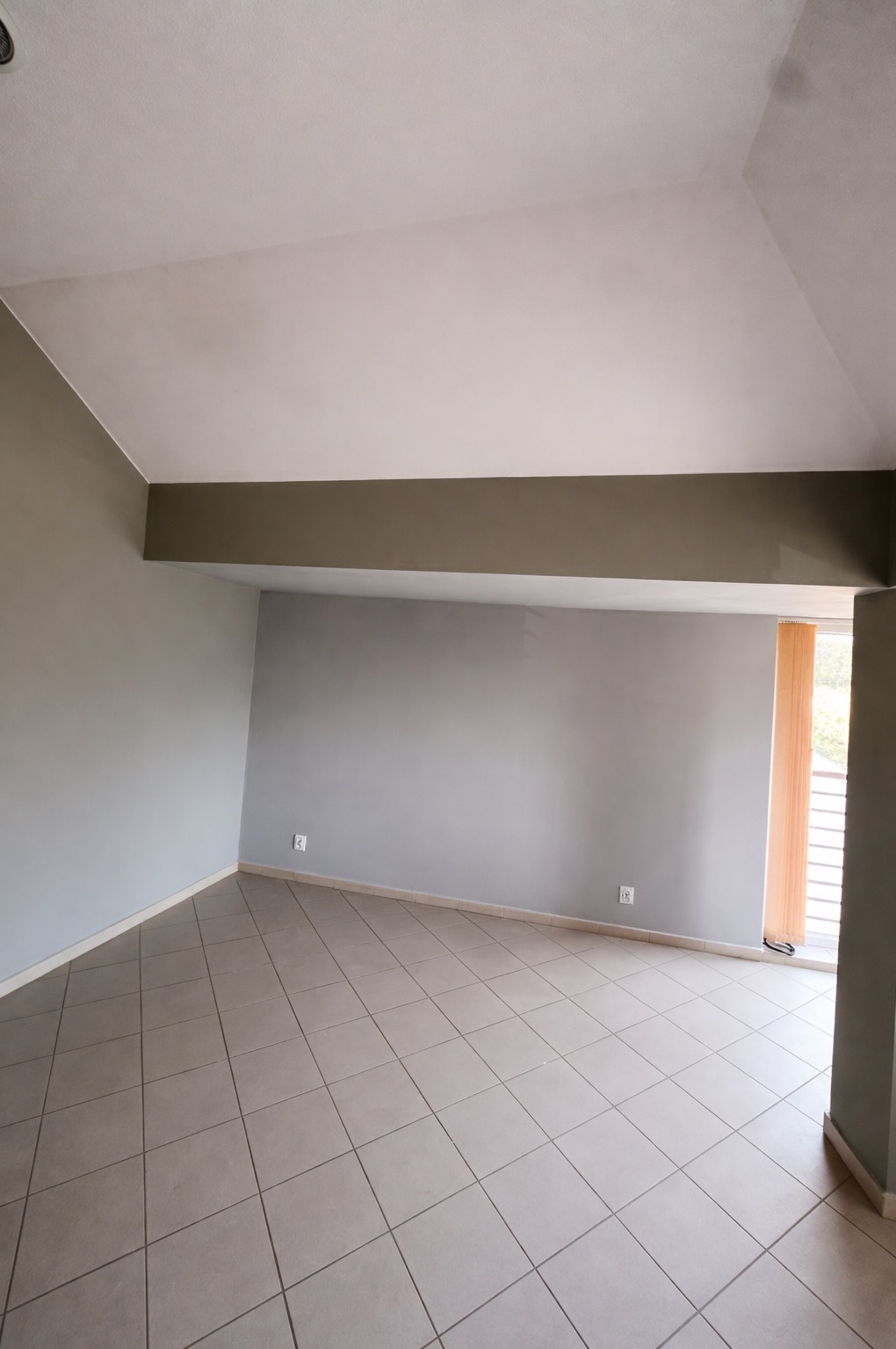 Pomalowane na szaro puste wnętrze z beżową podłogą z płytek, widoczne gniazdka elektryczne i fragment okna z żaluzjami. Świeżo po remoncie.