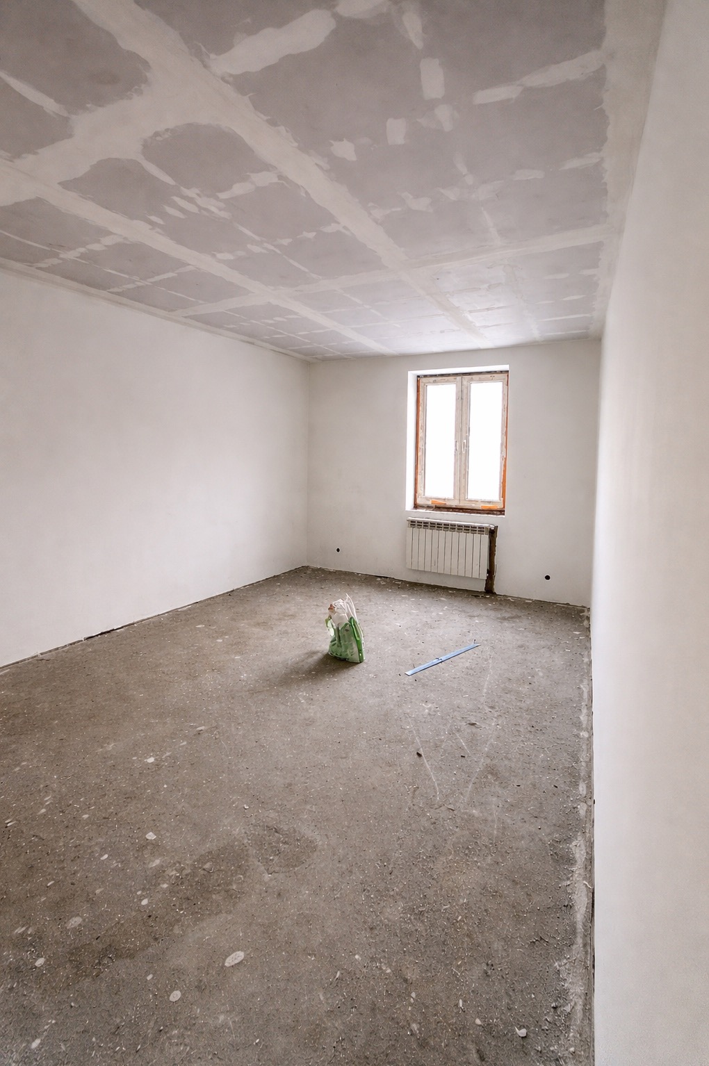 Pokój w trakcie remontu: surowy beton na podłodze, ściany przygotowane pod malowanie, częściowo wykończony sufit z widocznymi łączeniami płyt. Okno z grzejnikiem.