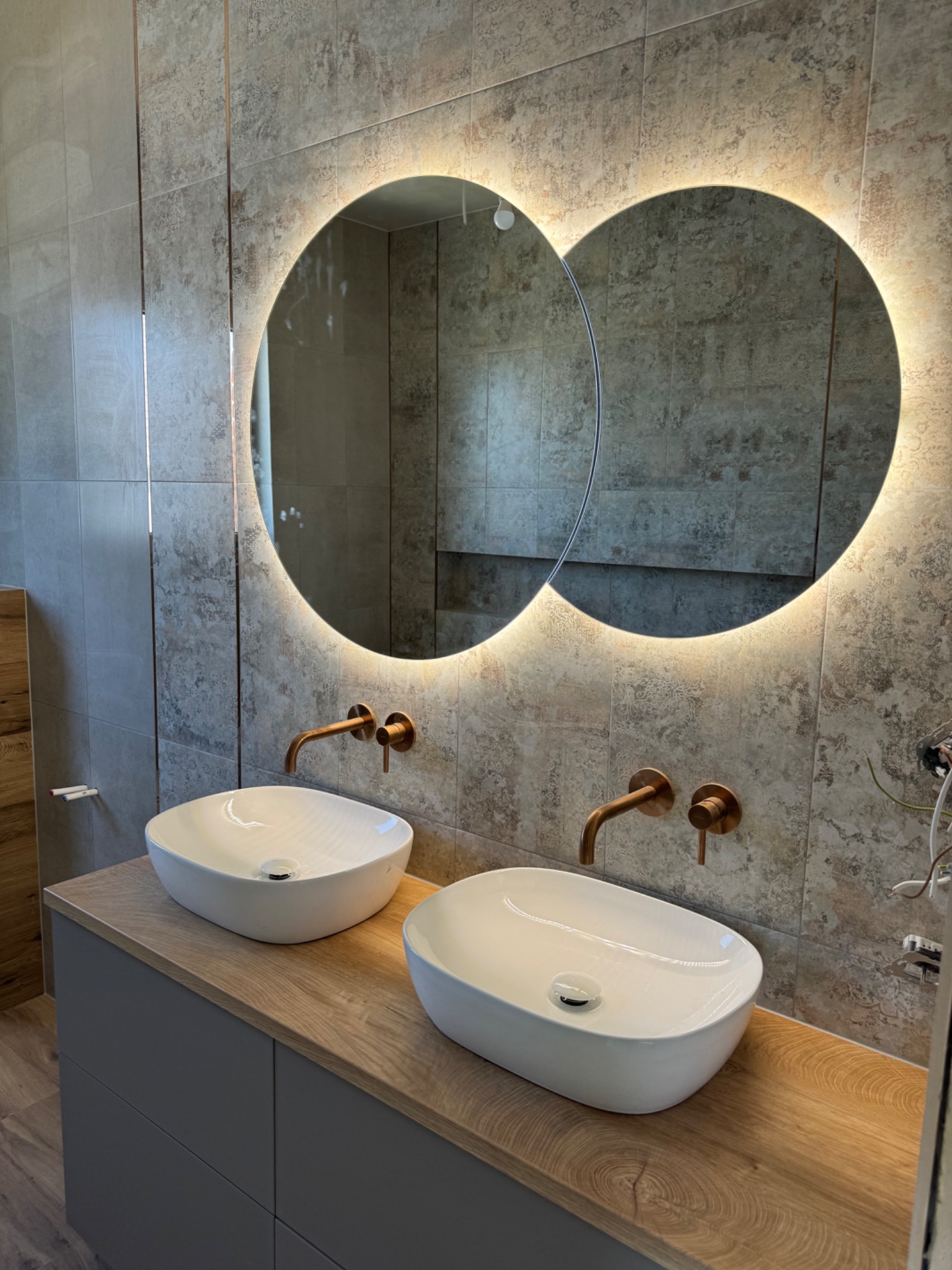Dwie białe umywalki nablatowe z miedzianymi kranami na drewnianym blacie, podświetlane okrągłe lustra i płytki w odcieniach szarości w łazience w Luboniu.
