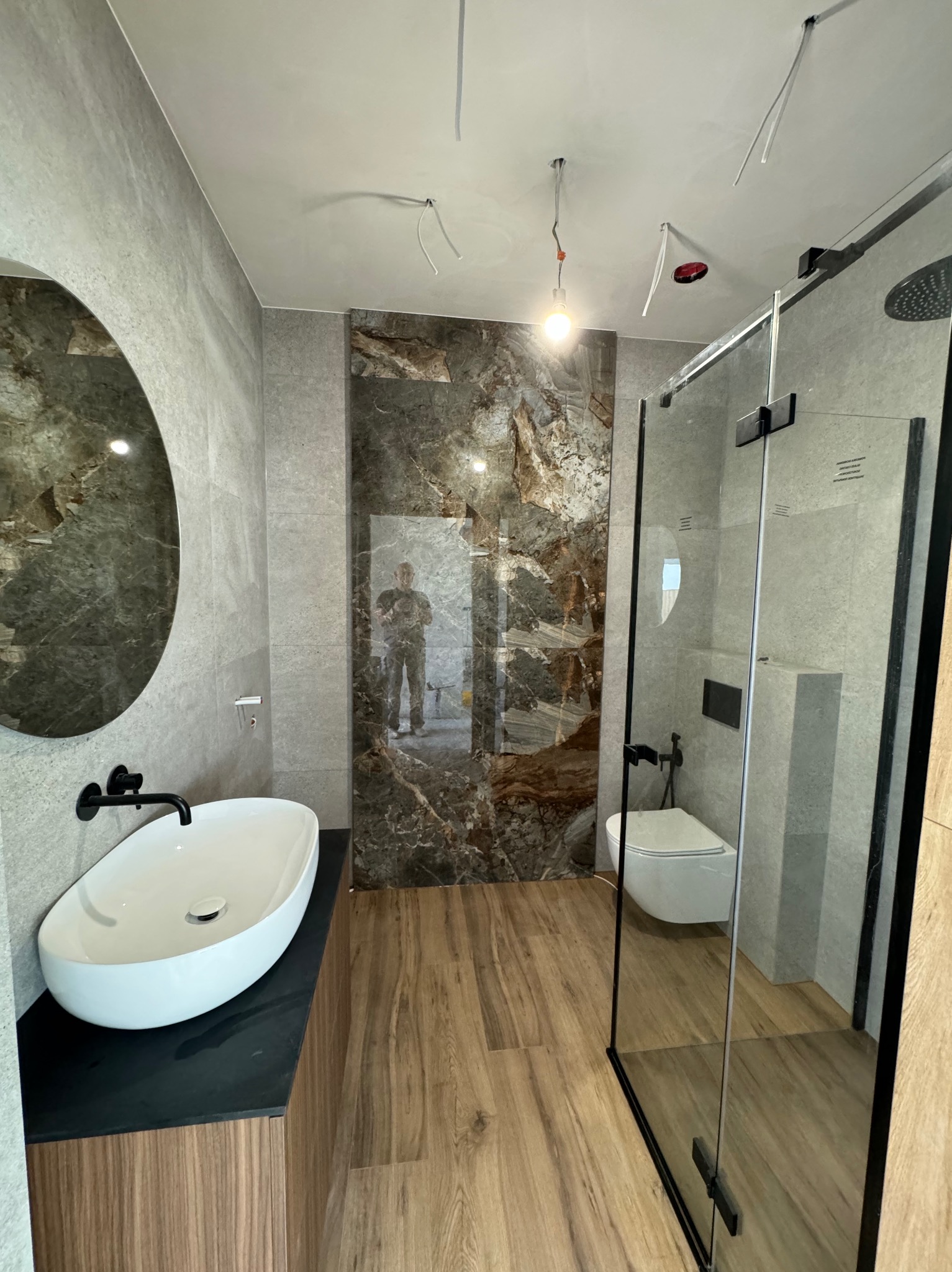 Nowoczesna łazienka z owalną umywalką nablatową, kabiną prysznicową typu walk-in z czarnymi profilami, płytkami imitującymi marmur na ścianie i drewnopodobną podłogą; widoczne niedokończone prace...
