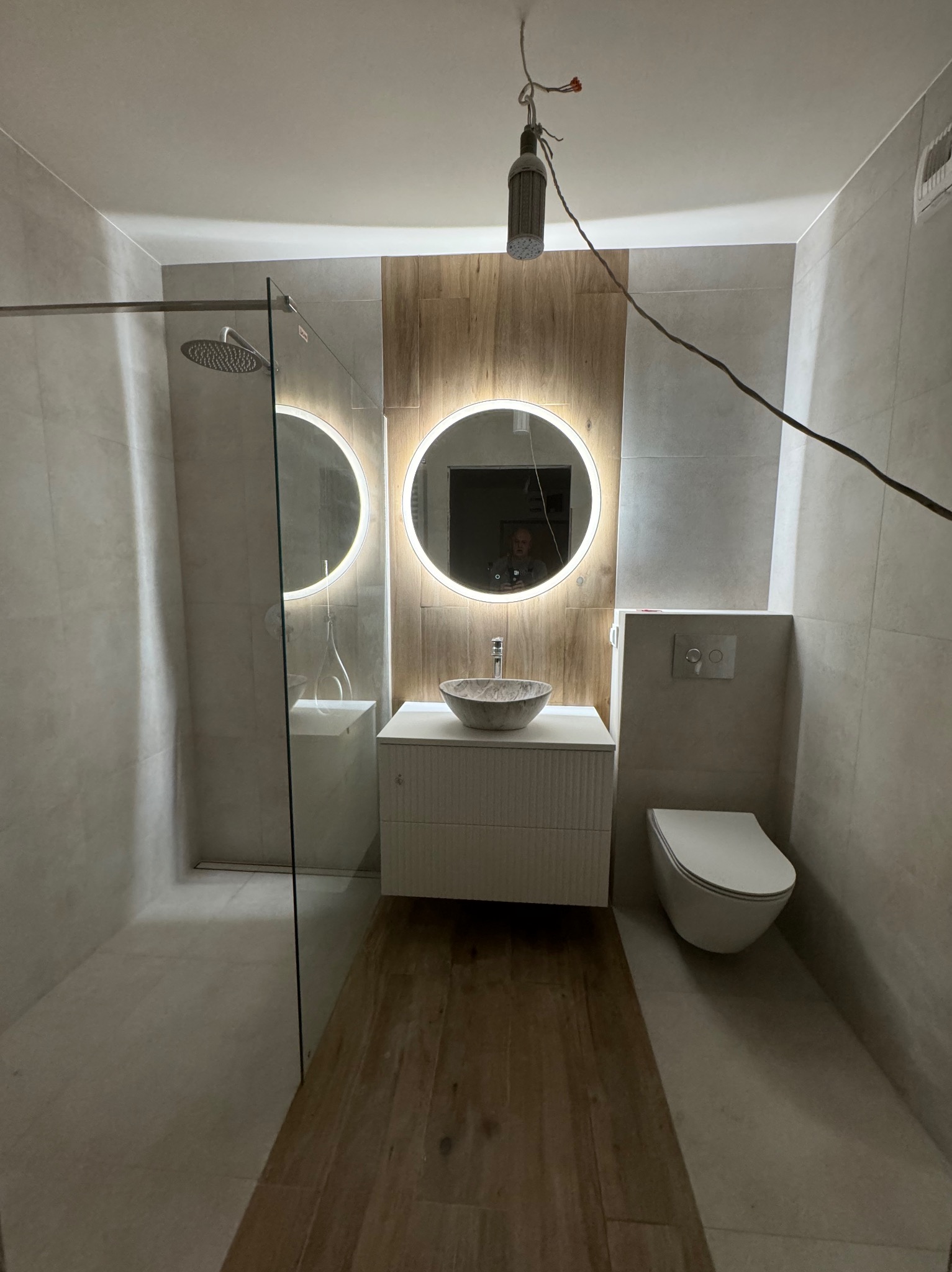 Nowoczesna łazienka w trakcie wykańczania: szara glazura, drewnopodobna podłoga, okrągłe lustra z podświetleniem, umywalka w stylu kamiennym, wisząca toaleta i kabina prysznicowa typu walk-in...
