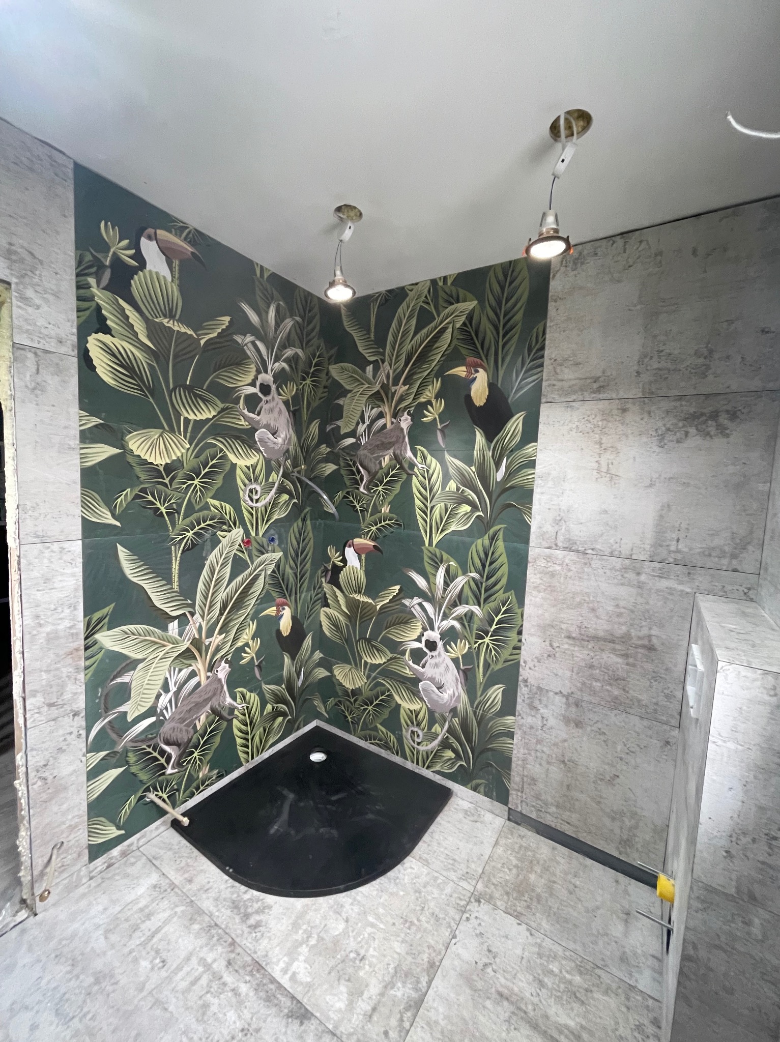 Wykończona łazienka z czarnym brodzikiem i płytkami imitującymi kamień, z fragmentem ściany pokrytej wzorem tropikalnej dżungli z tukanami i małpami, widoczne niedokończone elementy wykończeniowe.