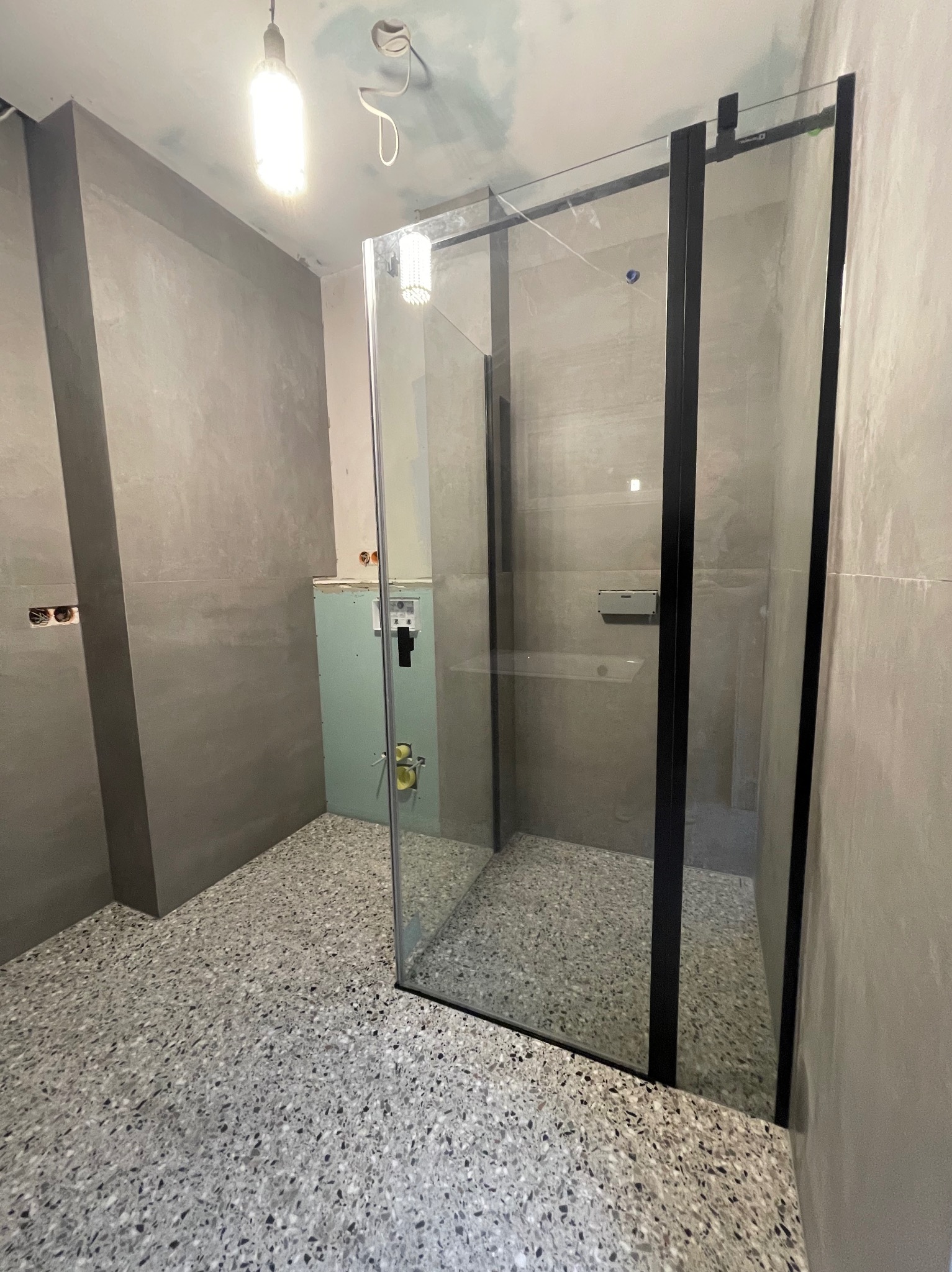 Nowoczesna kabina prysznicowa z czarnymi ramami w surowej łazience z szarymi ścianami i podłogą lastryko, widoczne niezakończone instalacje.