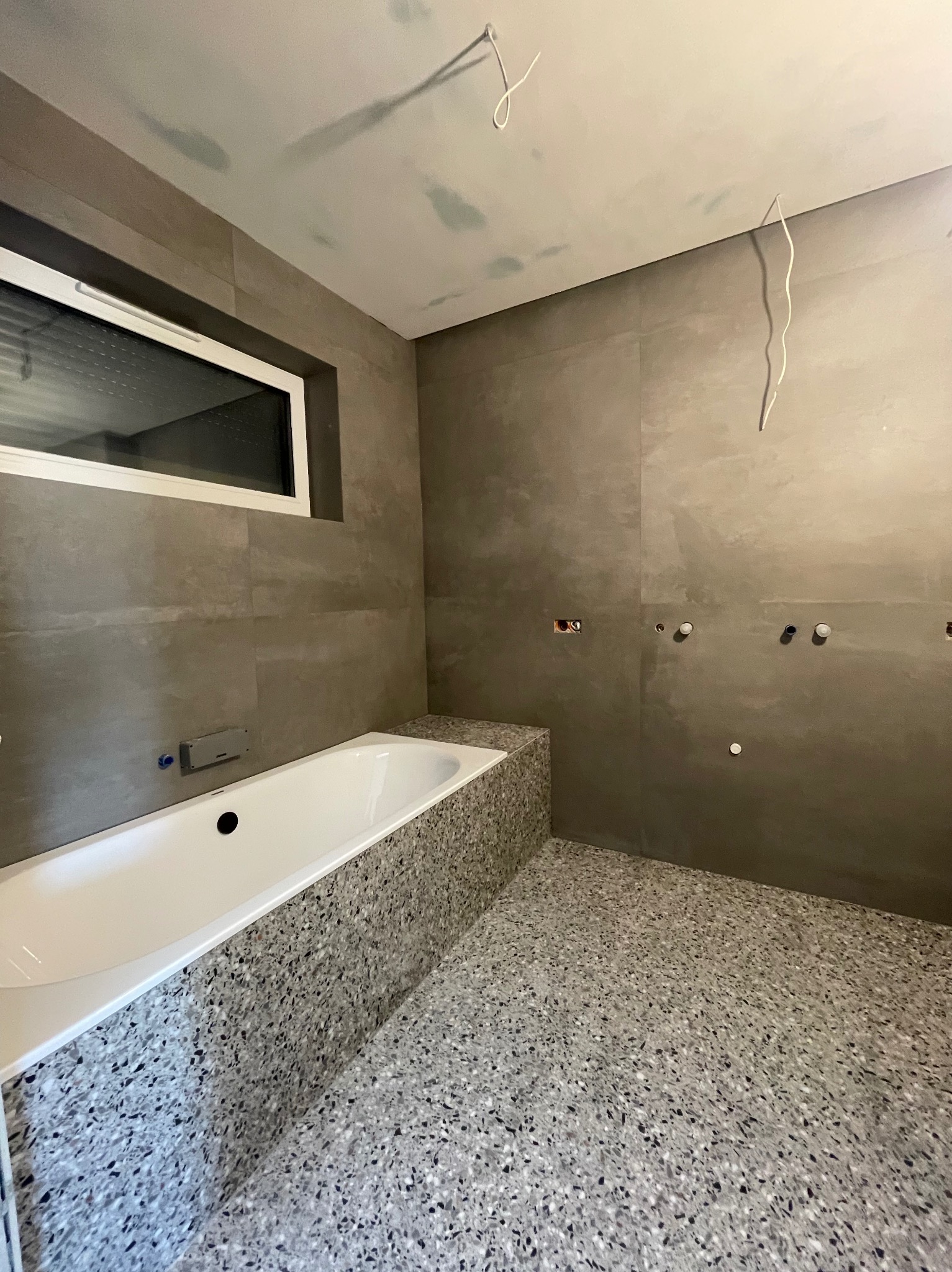 Surowa łazienka w trakcie wykończenia: szare płytki na ścianach, mozaika na obudowie wanny i podłodze, widoczne punkty instalacji wodnej i elektrycznej, sufit z niedomalowanymi fragmentami.