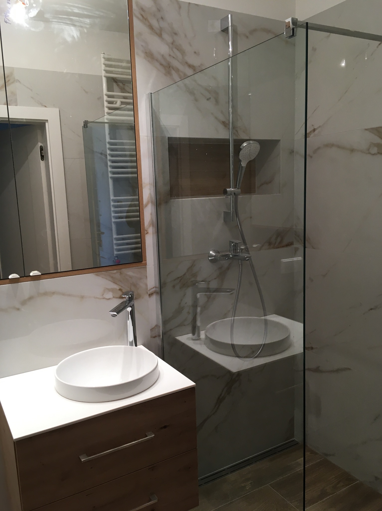 Nowoczesna łazienka z kabiną prysznicową typu walk-in, umywalką nablatową na drewnianej szafce z szufladami i lustrem w drewnianej ramie, wykończona płytkami imitującymi marmur.