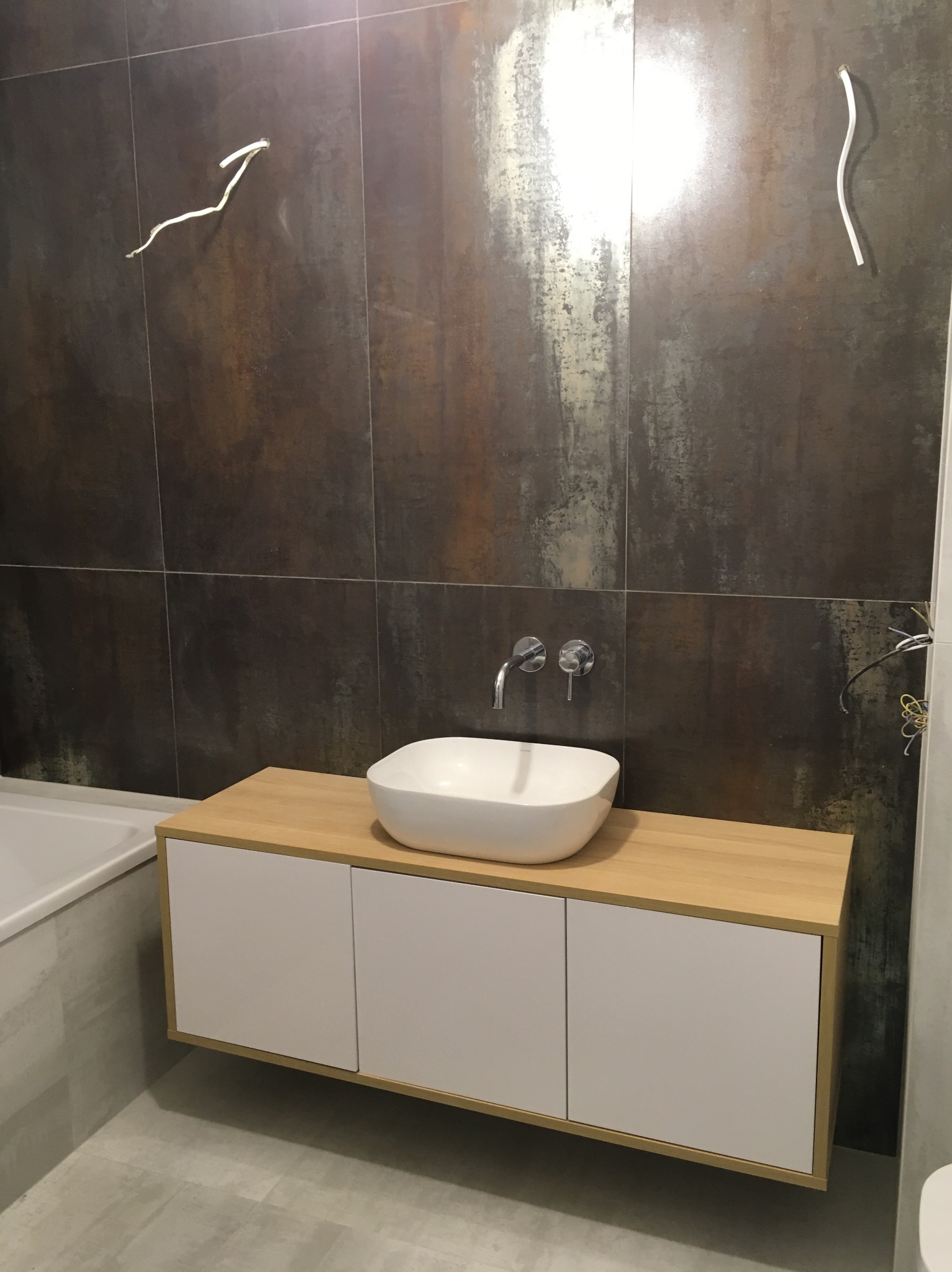 Nowoczesna łazienka z umywalką nablatową, szafką pod umywalką z białymi frontami i blatem z jasnego drewna, ściany wyłożone płytkami imitującymi rdzę, widoczne niedokończone podłączenie elektryczne.