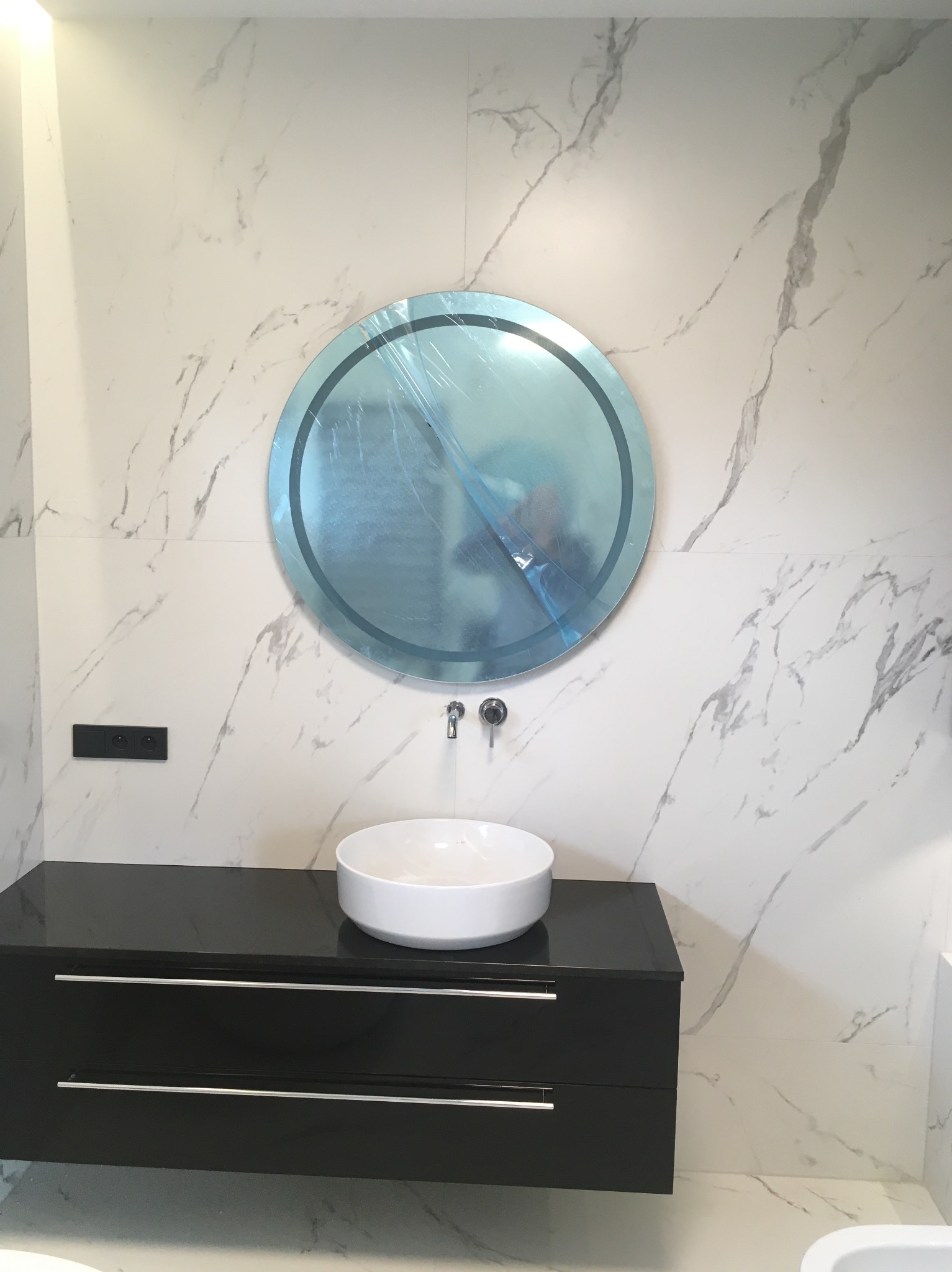 Nowoczesna łazienka z marmurową ścianą, okrągłym lustrem w folii ochronnej, białą umywalką nablatową i czarną szafką pod umywalką z chromowanymi uchwytami, widoczny czarny włącznik elektryczny.