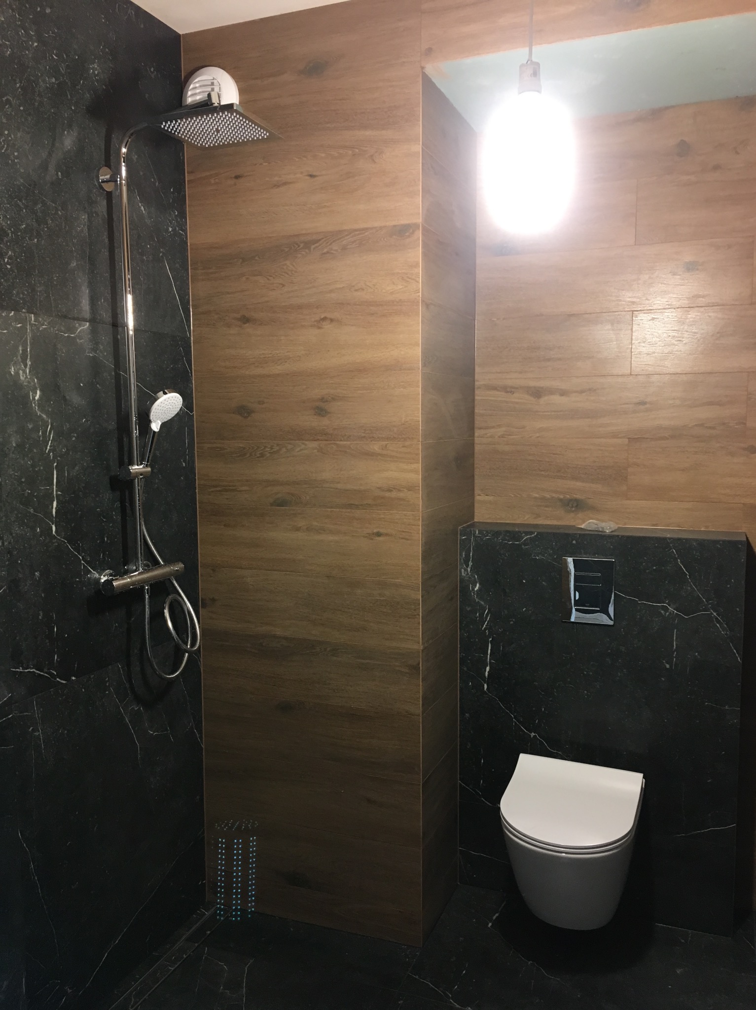 Nowoczesna łazienka z czarnymi marmurowymi płytkami i drewnopodobnymi panelami, wyposażona w deszczownicę, toaletę podwieszaną i oświetlenie sufitowe.