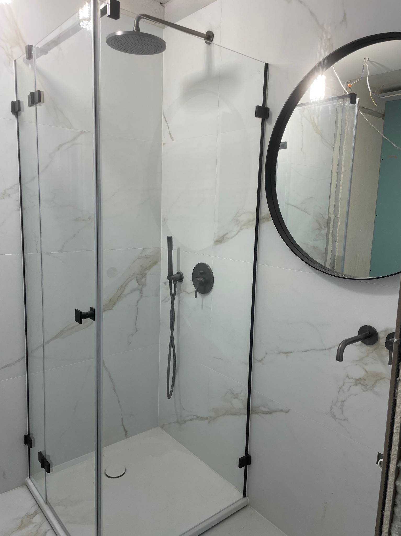 Nowoczesna kabina prysznicowa z czarnymi okuciami i armaturą na tle białych płytek imitujących marmur oraz okrągłe lustro z czarną ramą, widoczne elementy instalacji elektrycznej w odbiciu lustra.