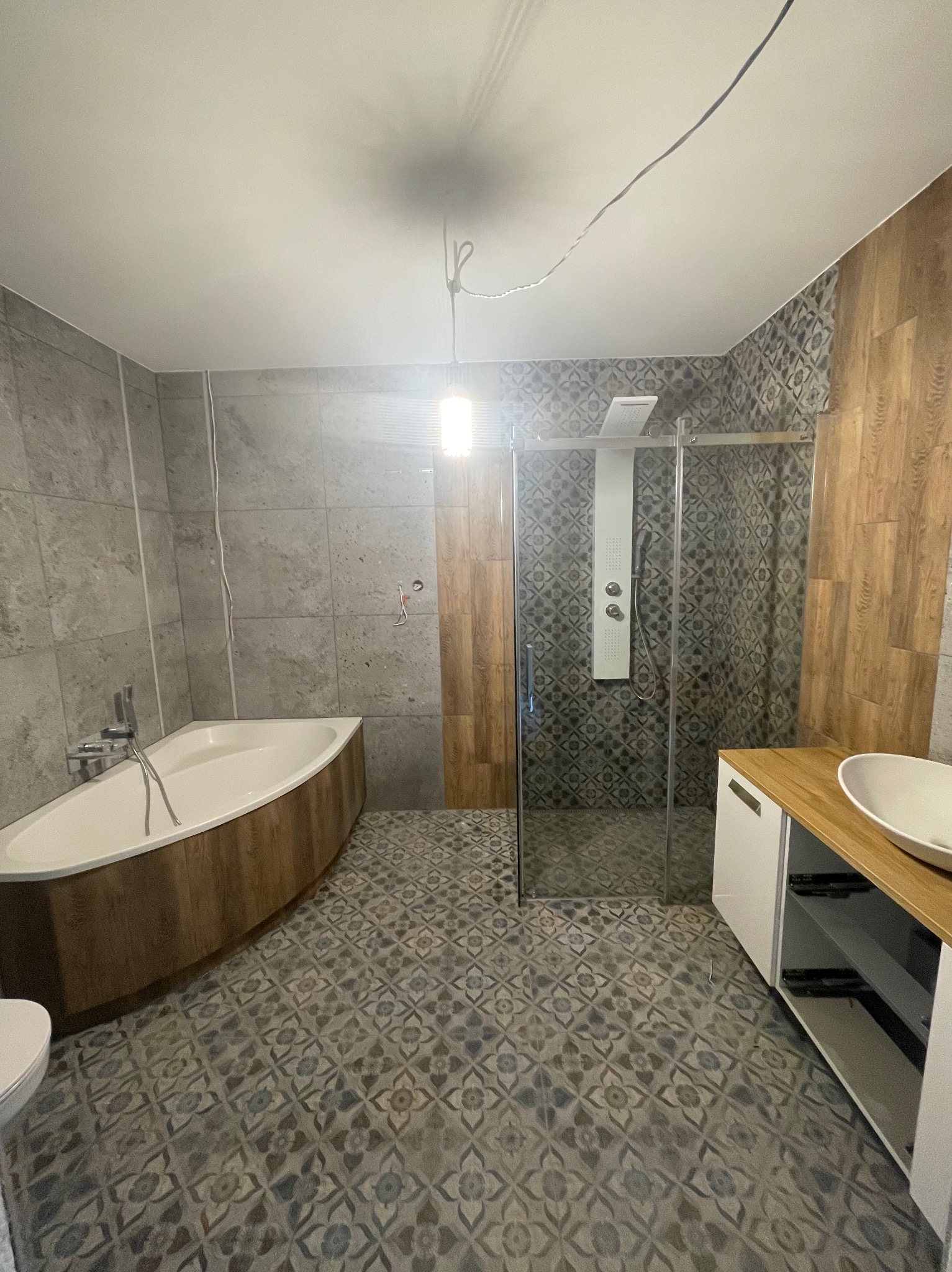 Nowoczesna łazienka z narożną wanną obudowaną drewnem, kabiną prysznicową typu walk-in z panelem natryskowym i umywalką nablatową na szafce z drewnianym blatem, wykończona płytkami w odcieniach...