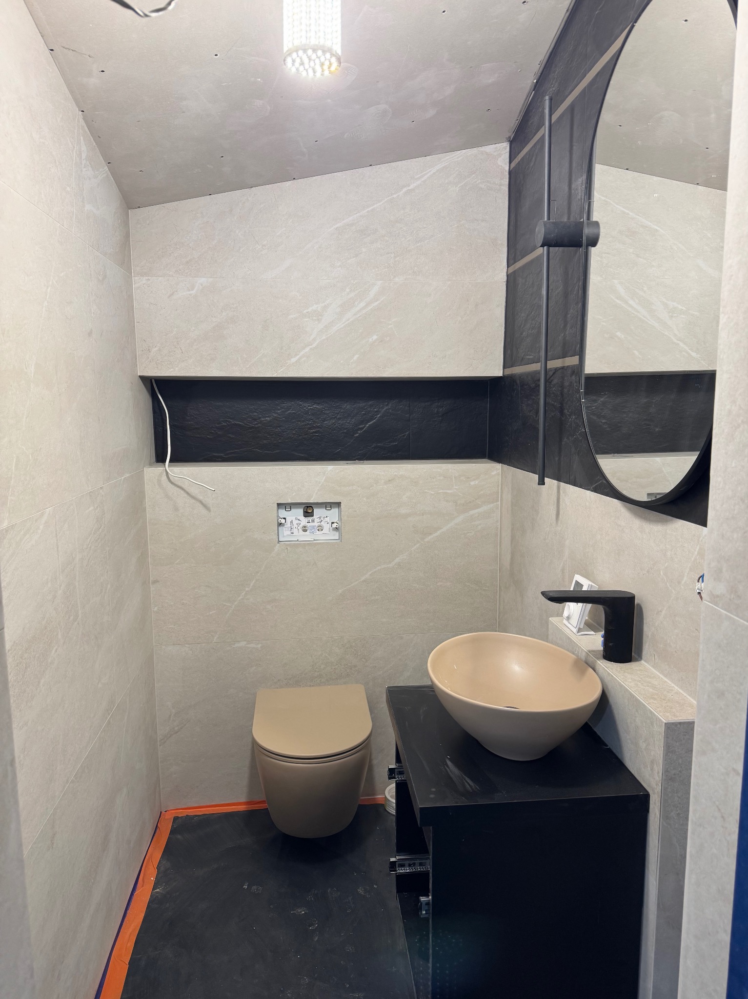 Nowoczesna łazienka w trakcie wykańczania: beżowe płytki imitujące marmur, czarne akcenty, okrągłe lustro i beżowa umywalka nablatowa. Widoczne elementy instalacji.