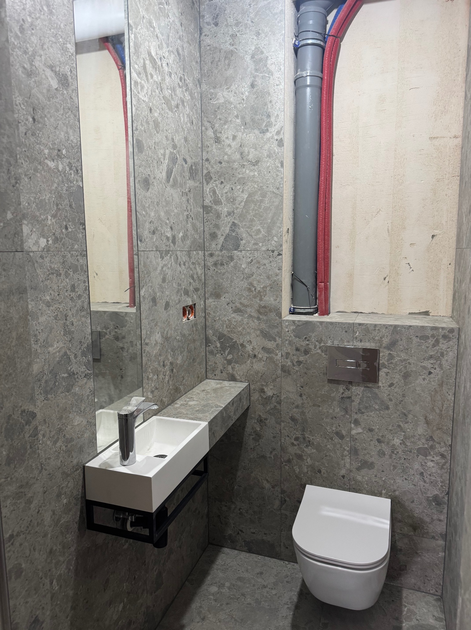Szara łazienka z marmurowymi płytkami, podwieszana umywalka z czarnym stelażem, toaleta i rury w ścianie. Widoczne niedokończone prace przy rurach.