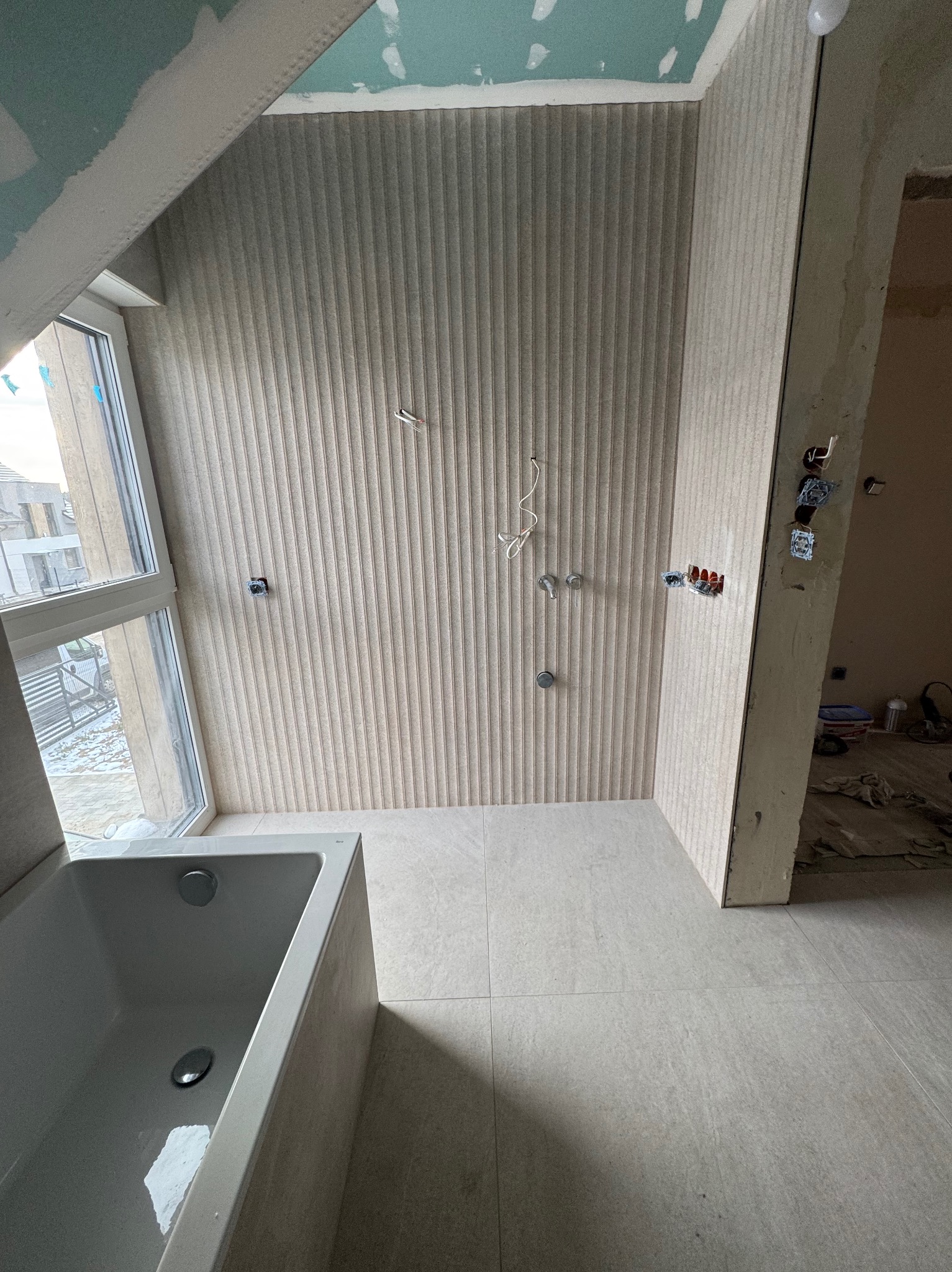 Łazienka w trakcie wykończenia: ułożone płytki ścienne z fakturą, przygotowane instalacje, widoczna wanna i okno z widokiem na zewnątrz.