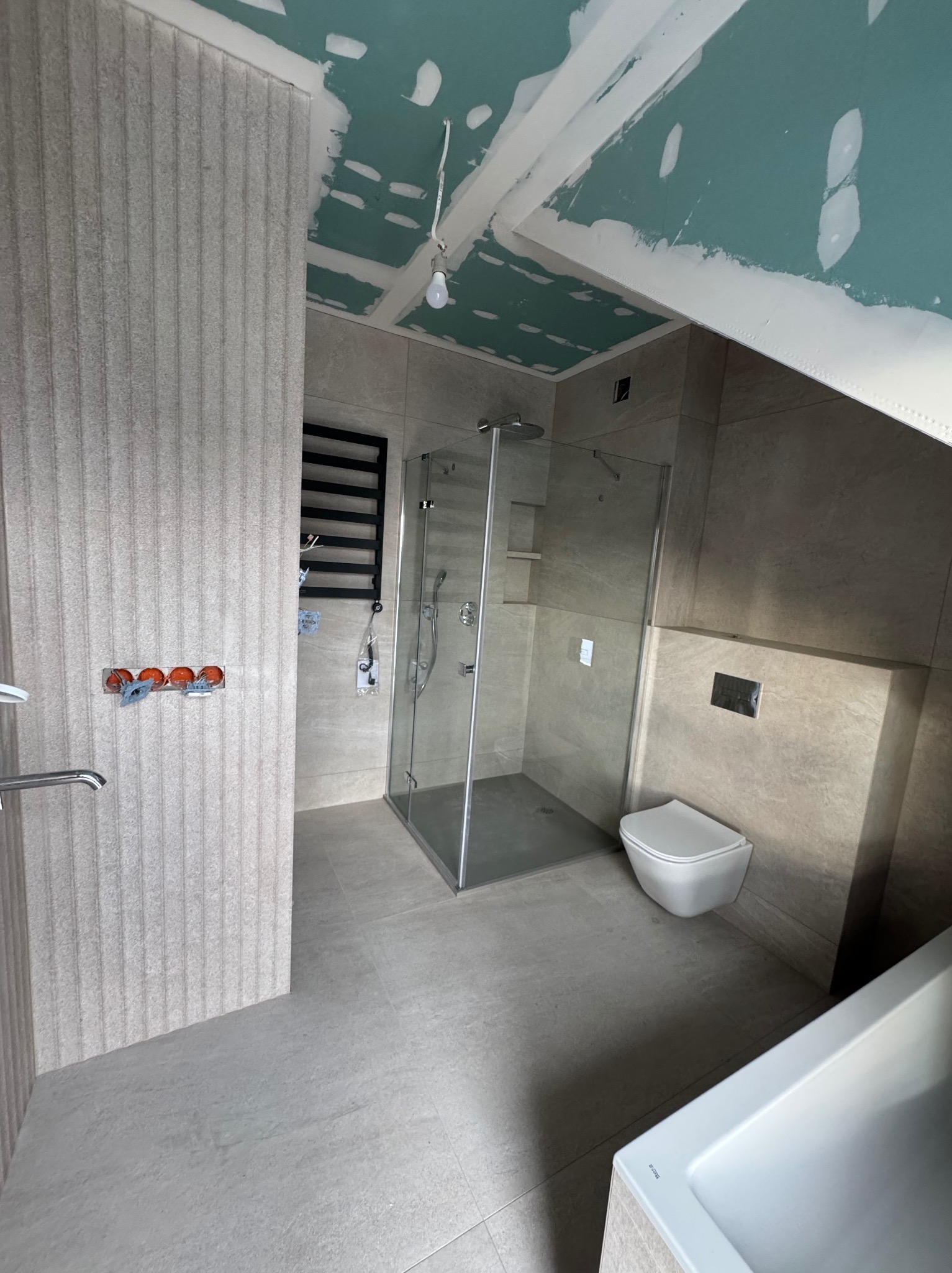 Nowoczesna łazienka w trakcie wykończenia: kabina prysznicowa, podwieszana toaleta, grzejnik drabinkowy na ścianie, widoczne instalacje elektryczne i sufit z płyt gipsowych.