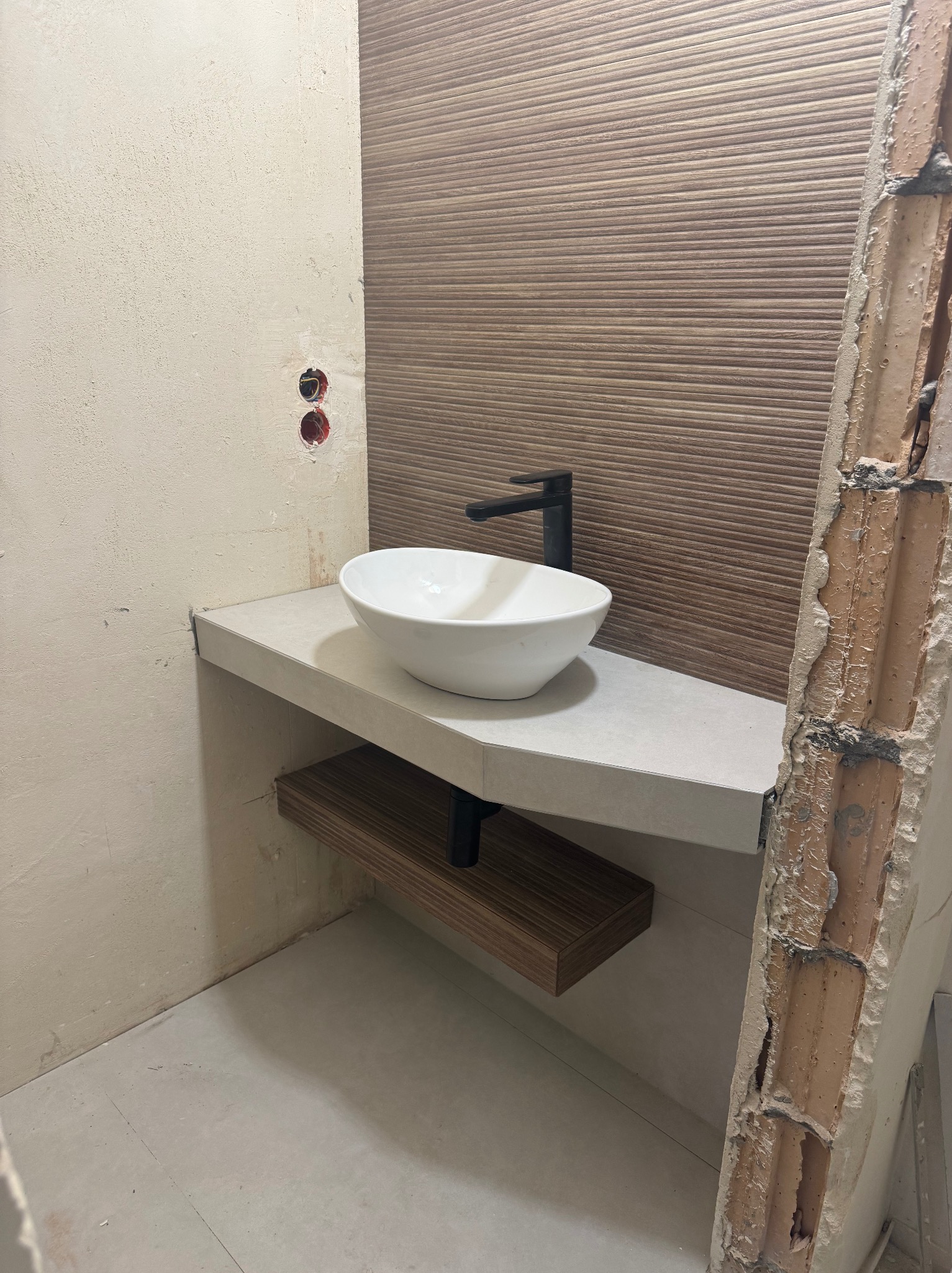Biała umywalka nablatowa z czarną armaturą na szarym blacie z półką w łazience, ściana wyłożona brązowymi płytkami. Fragment ściany w stanie surowym.