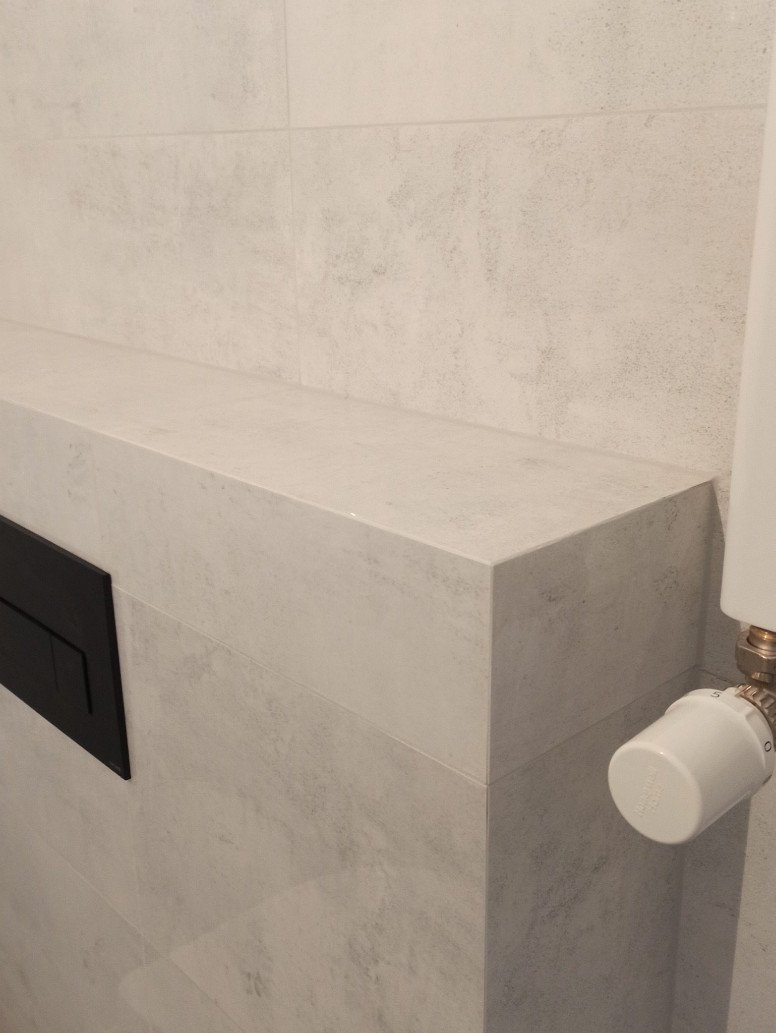 Wykończona ściana w łazience z szarymi płytkami imitującymi kamień, czarny panel sterowania spłuczką i biały termostat grzejnika.