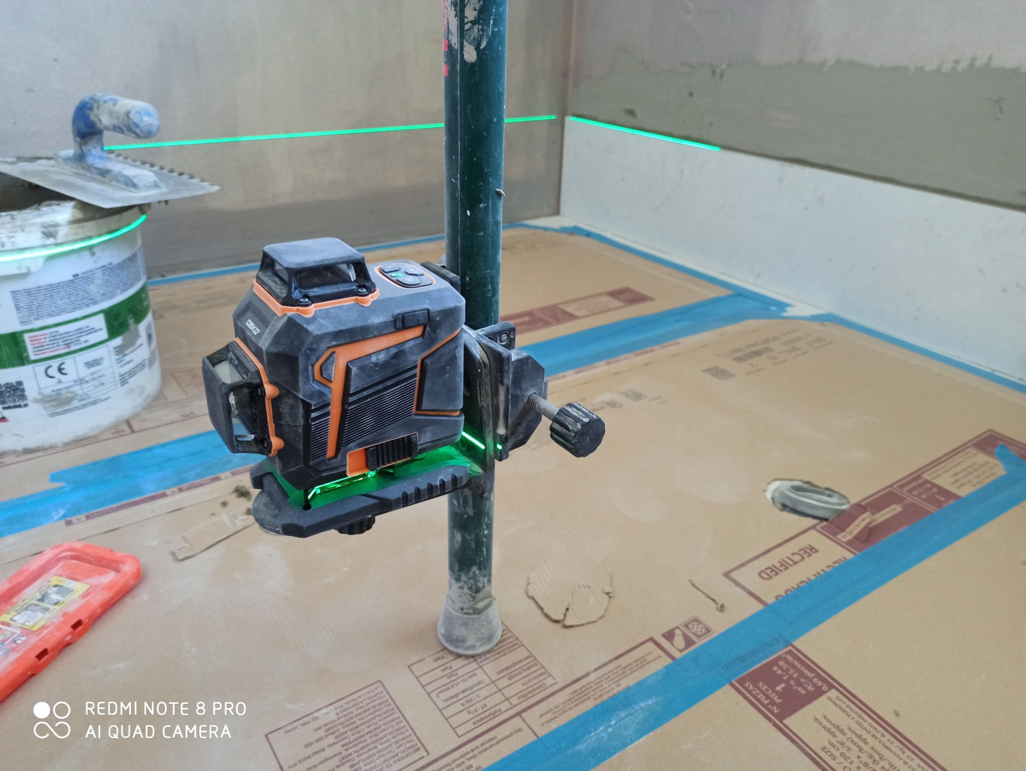 Poziomica laserowa DEKO, ustawiona na metalowej rurze, wyznacza poziom na ścianie w trakcie prac wykończeniowych; obok wiadro z zaprawą i kielnia.