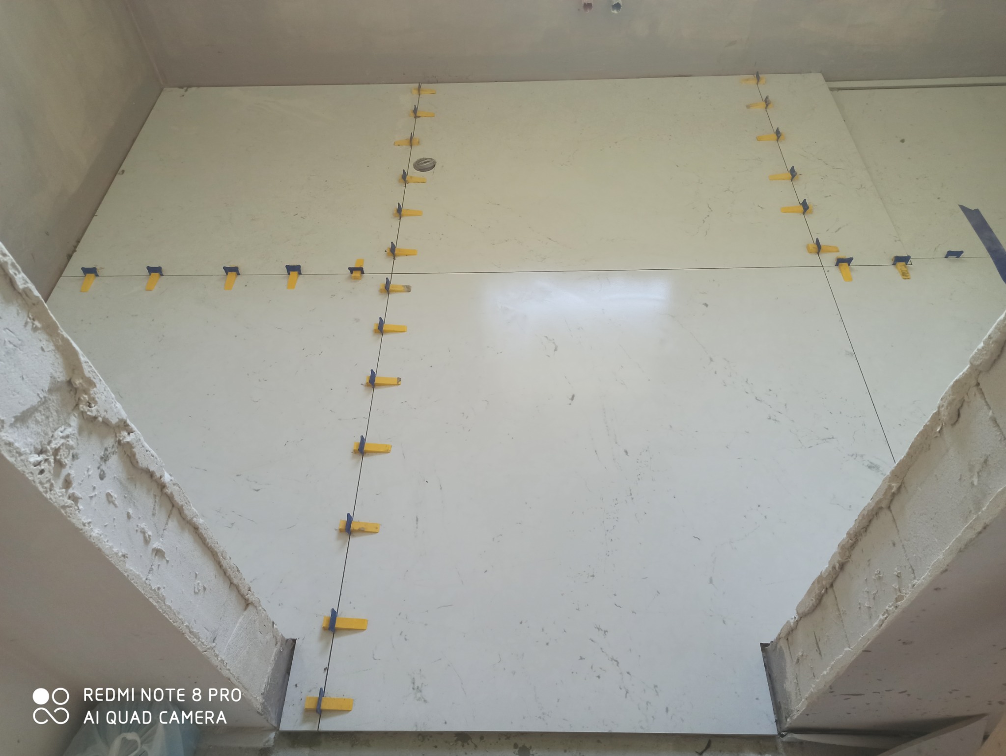 Białe płytki imitujące marmur, ułożone na ścianie z użyciem poziomicy i żółtych klipsów dystansowych, widoczne surowe wykończenie ściany.
