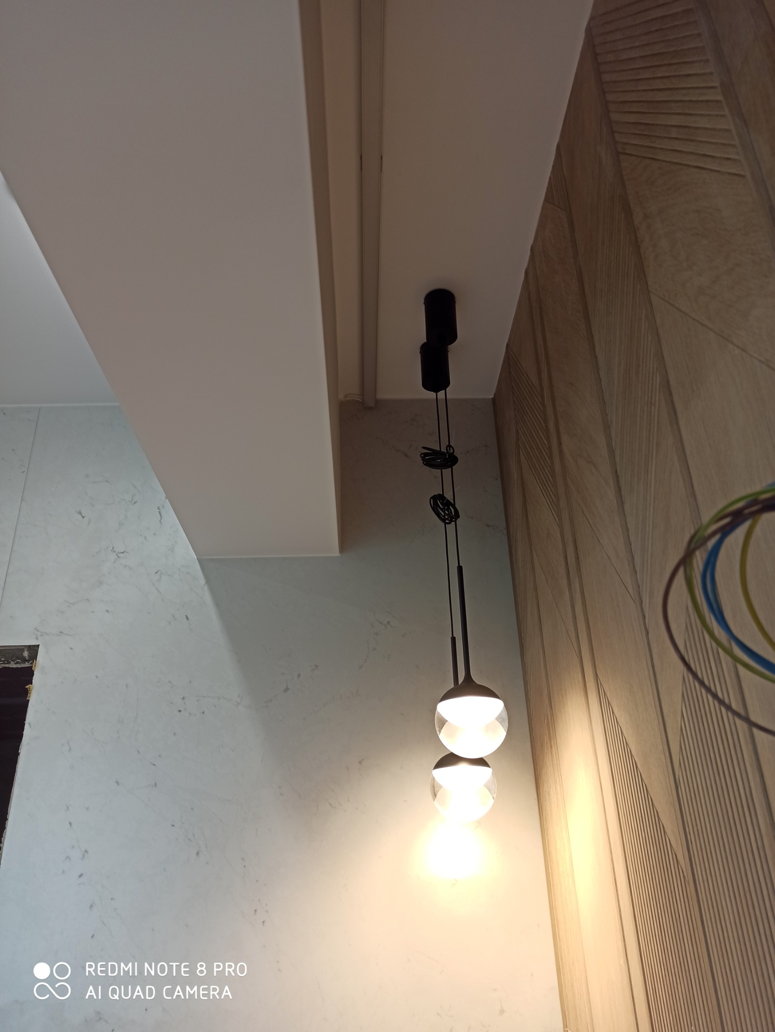 Nowoczesna lampa wisząca z dwoma kulistymi kloszami, zamontowana na suficie w pomieszczeniu z wykończonymi ścianami: jedną w marmurze, drugą w drewnianej okładzinie.