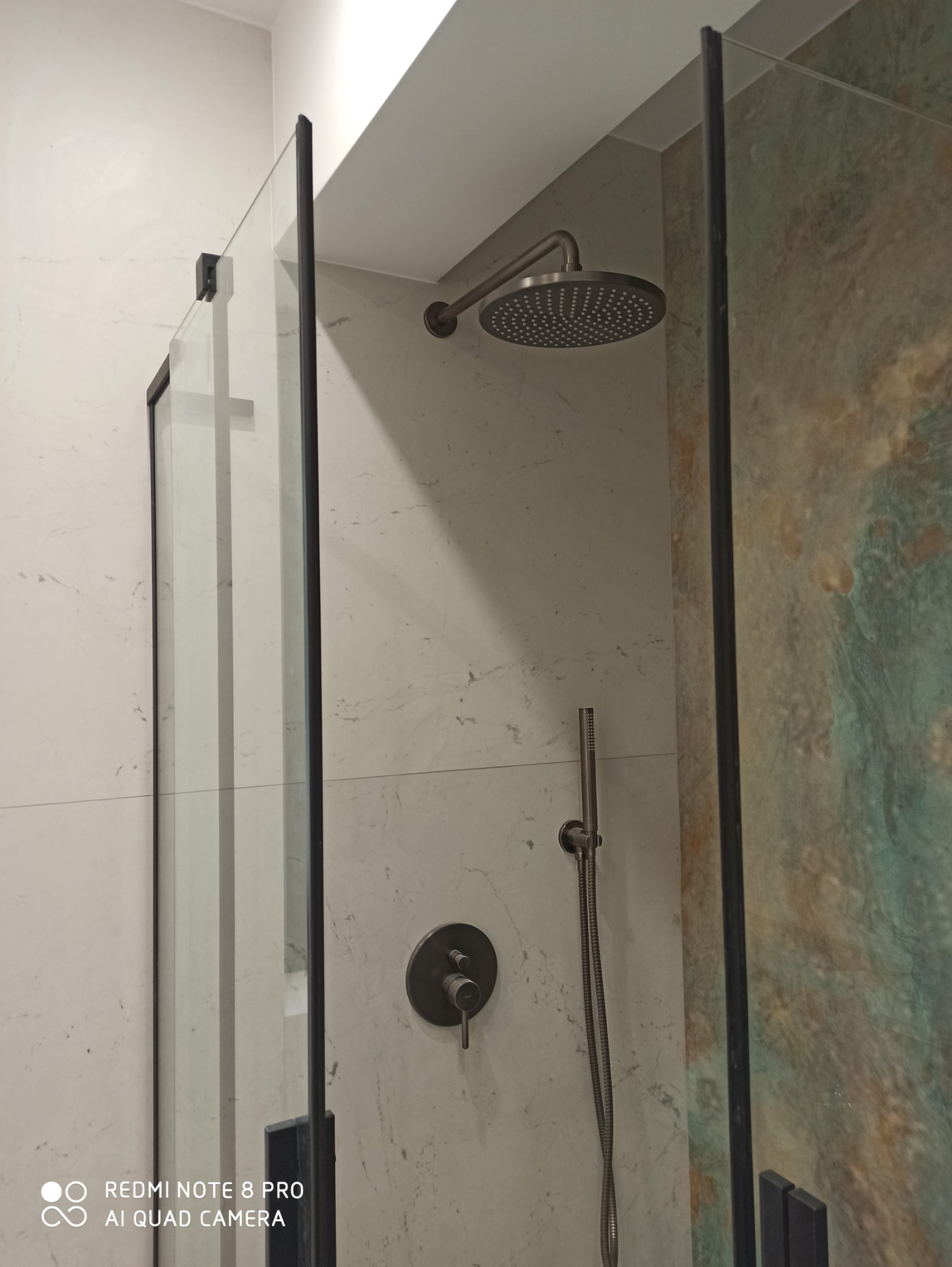 Nowoczesna kabina prysznicowa z czarnymi ramami, deszczownica i bateria w kolorze szczotkowanego metalu na tle marmurowej ściany i panelu z efektem akwareli.