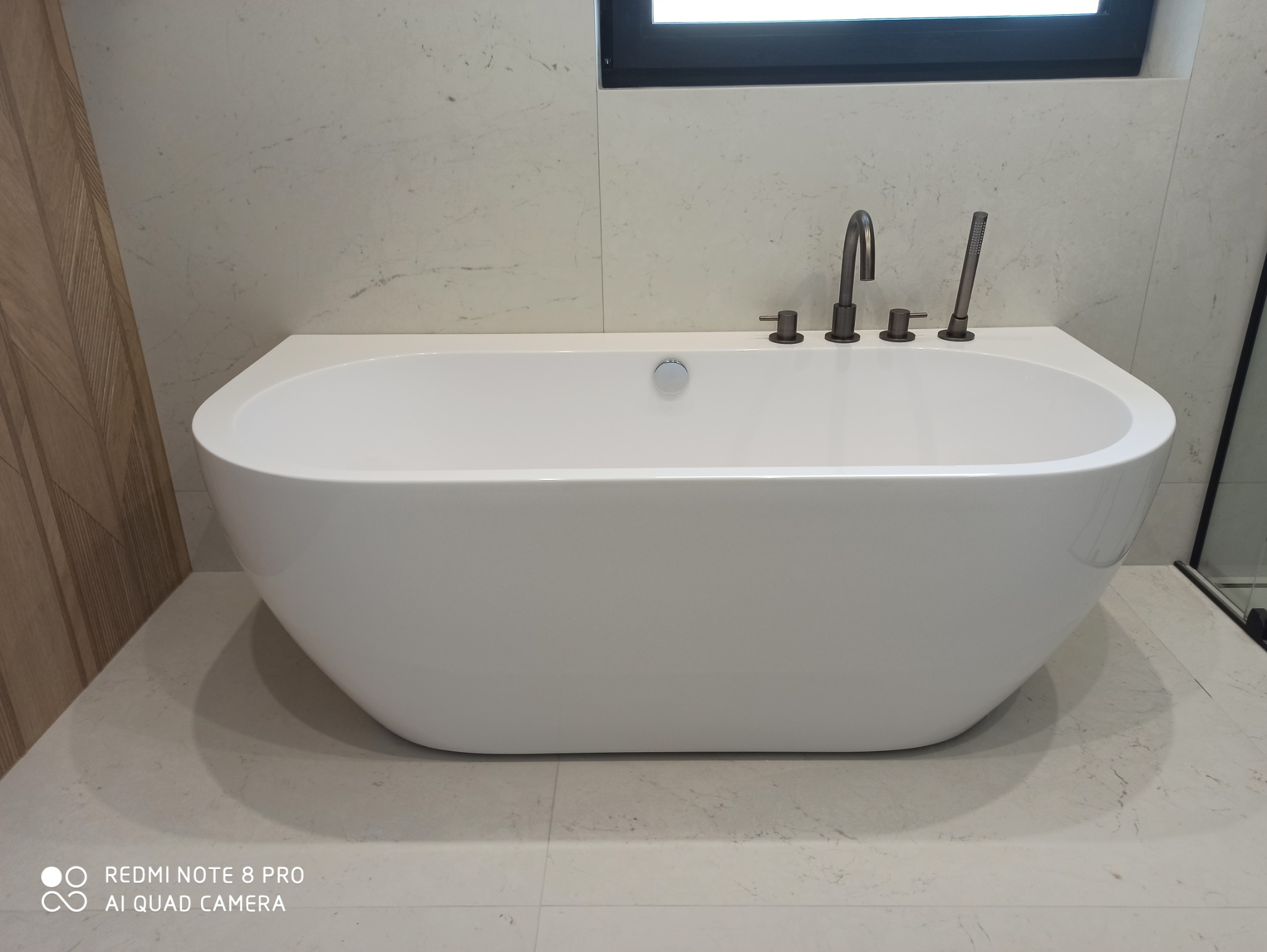 Nowoczesna, wolnostojąca wanna z ciemną armaturą na tle marmurowych płytek w łazience z oknem.